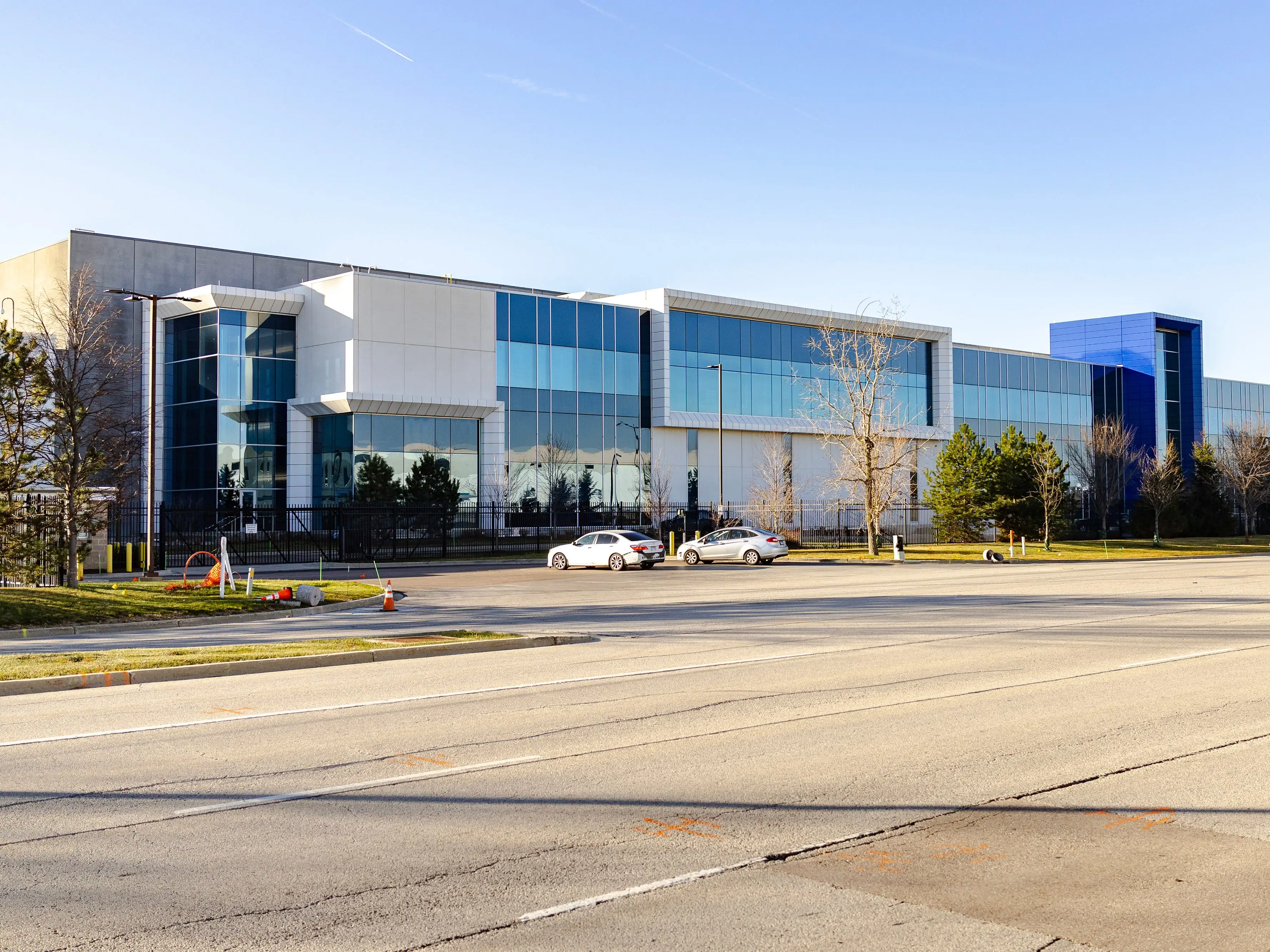 Modern glass office building stands behind a wide road with parked cars and trees under a clear sky.