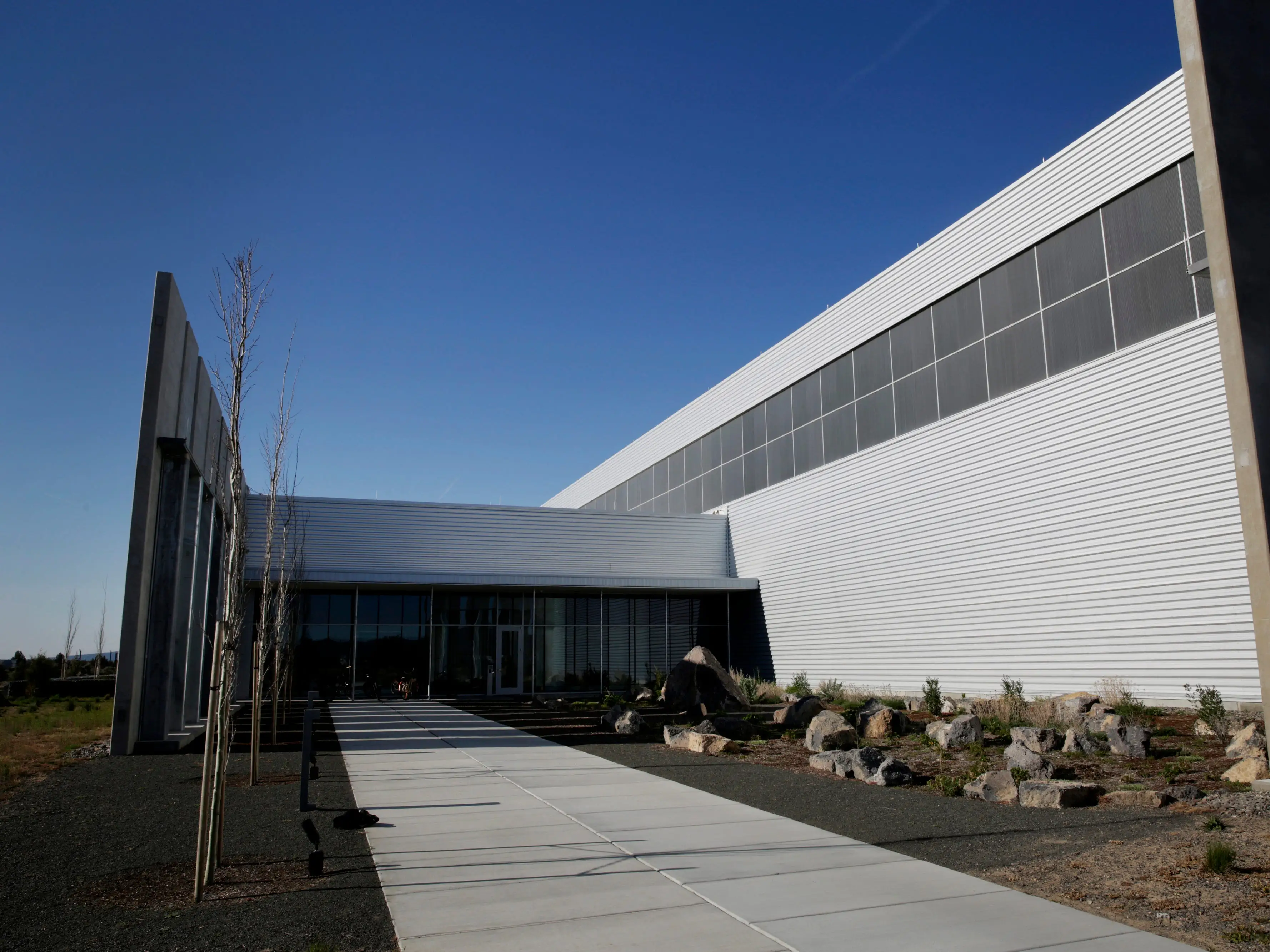 Modern white industrial building with large dark windows, concrete walkway, rocks, and sparse landscaping under a clear blue sky.