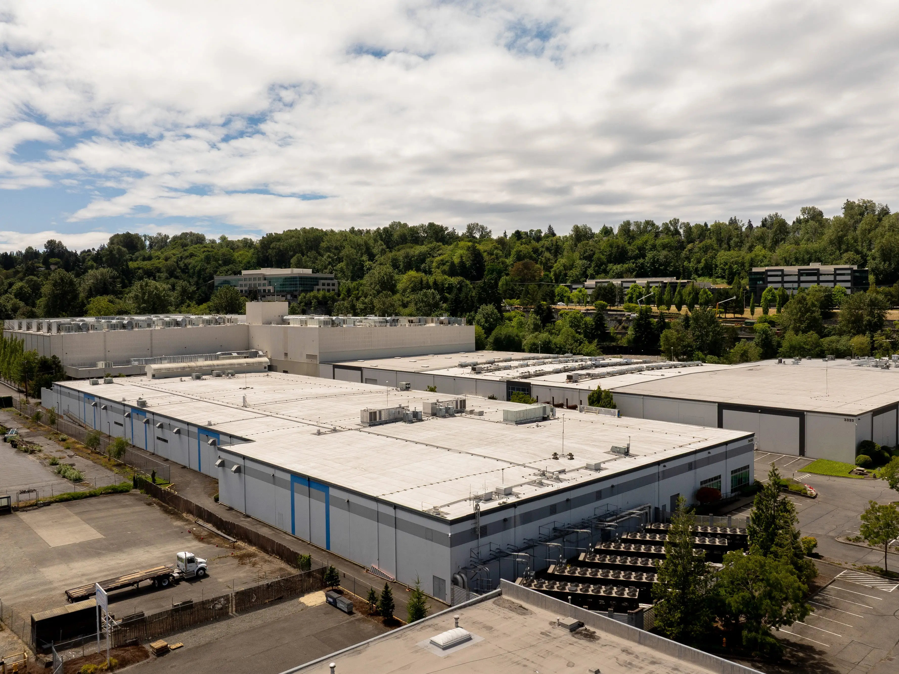 Large industrial warehouse complex with flat roofs surrounded by trees and roads under a cloudy sky.