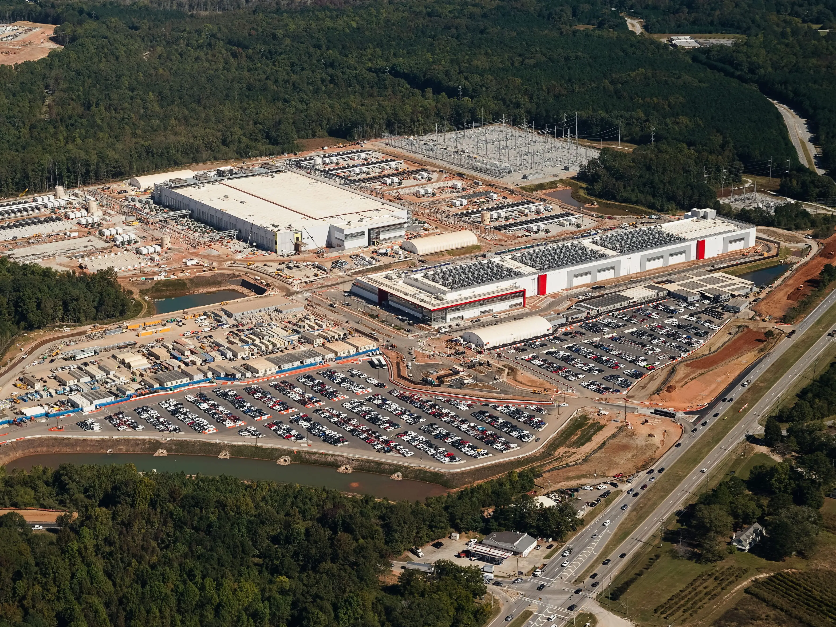 Aerial view of a large industrial complex under construction surrounded by parking lots, roads, forest, and power infrastructure.