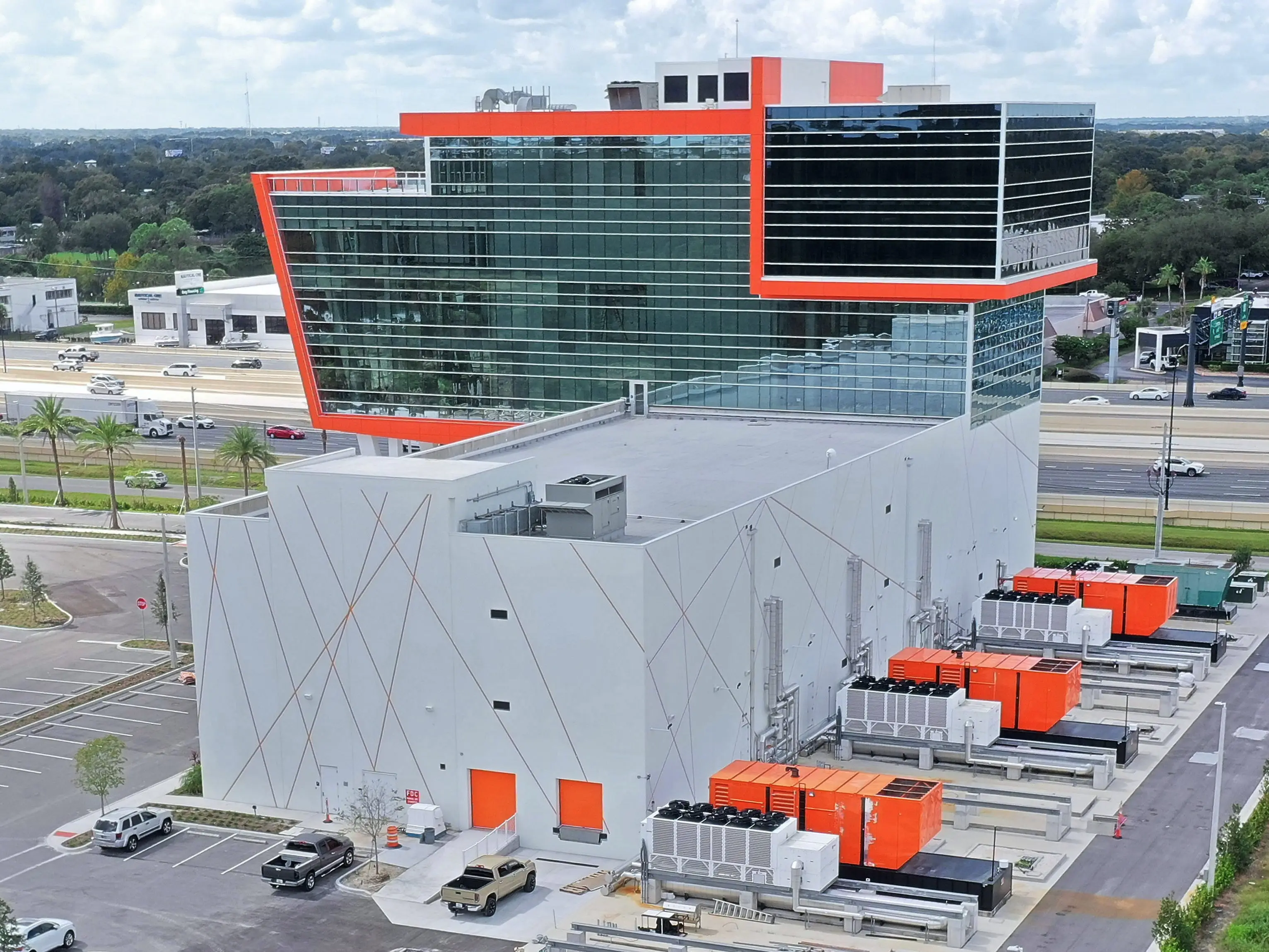 Aerial view of a modern glass and concrete facility with orange accents beside a multi-lane highway.