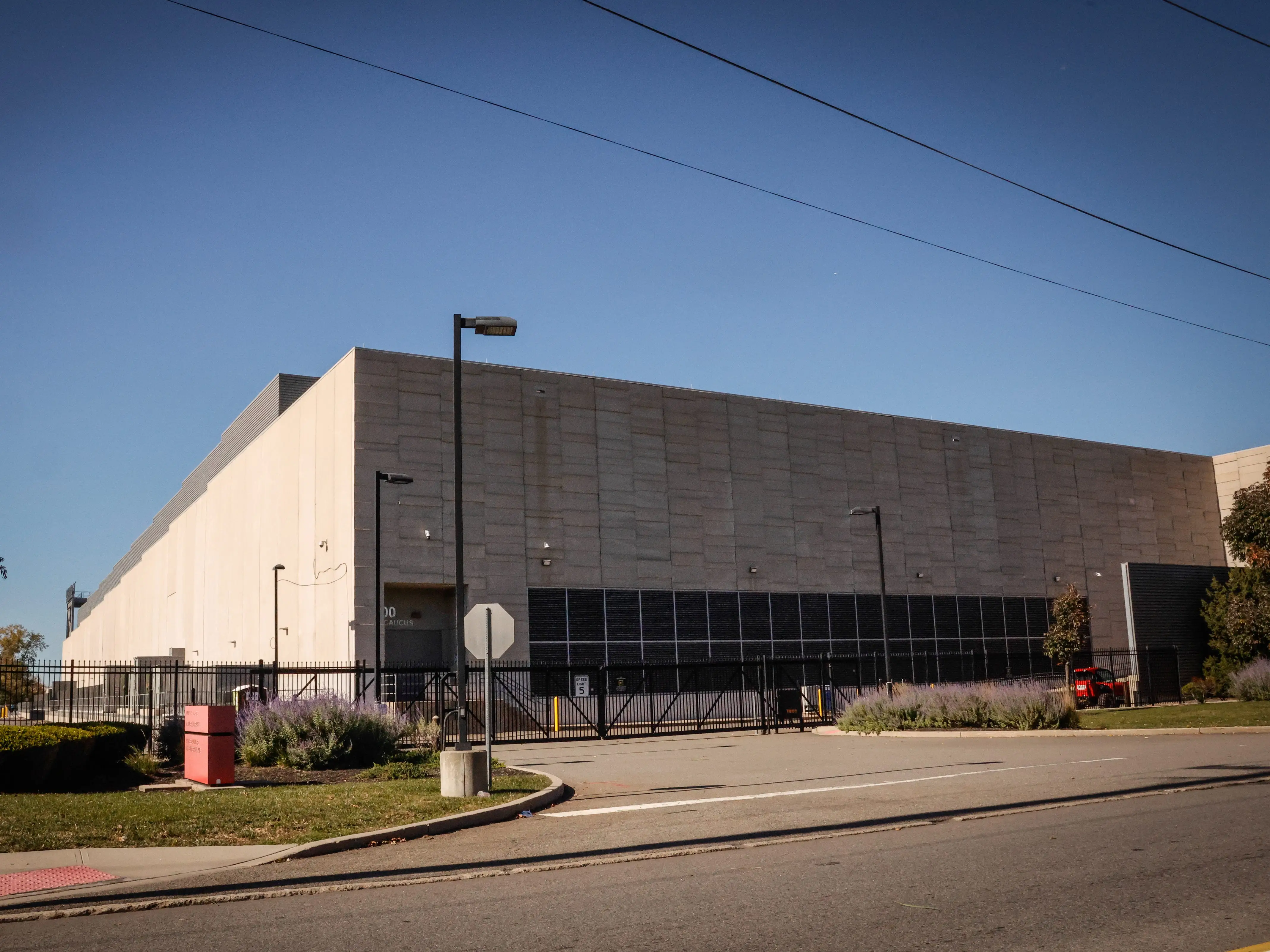 Large windowless concrete building sits behind a black fence along a quiet street under a clear blue sky.