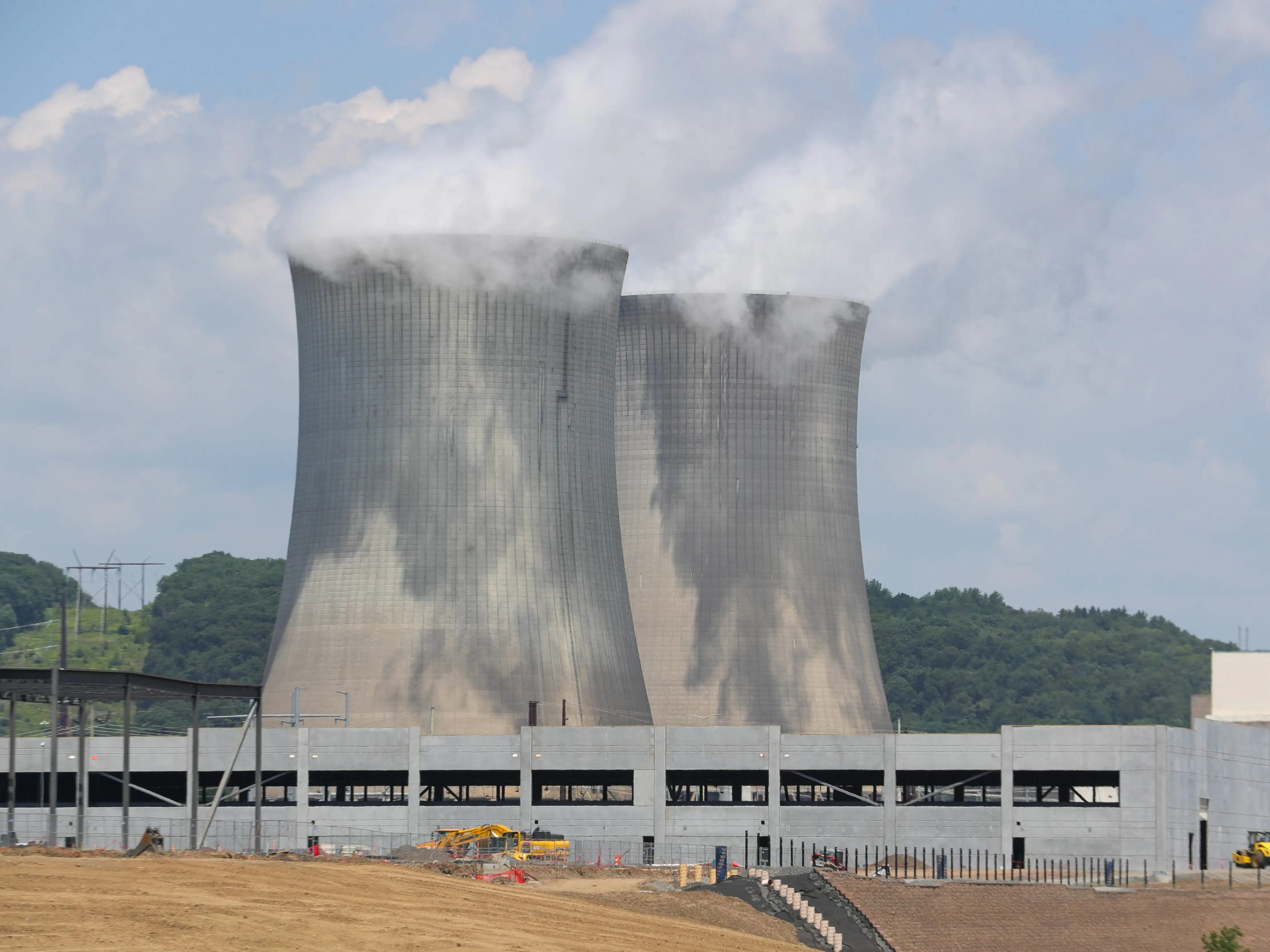 Two large cooling towers release steam behind an industrial facility construction site under a partly cloudy sky.