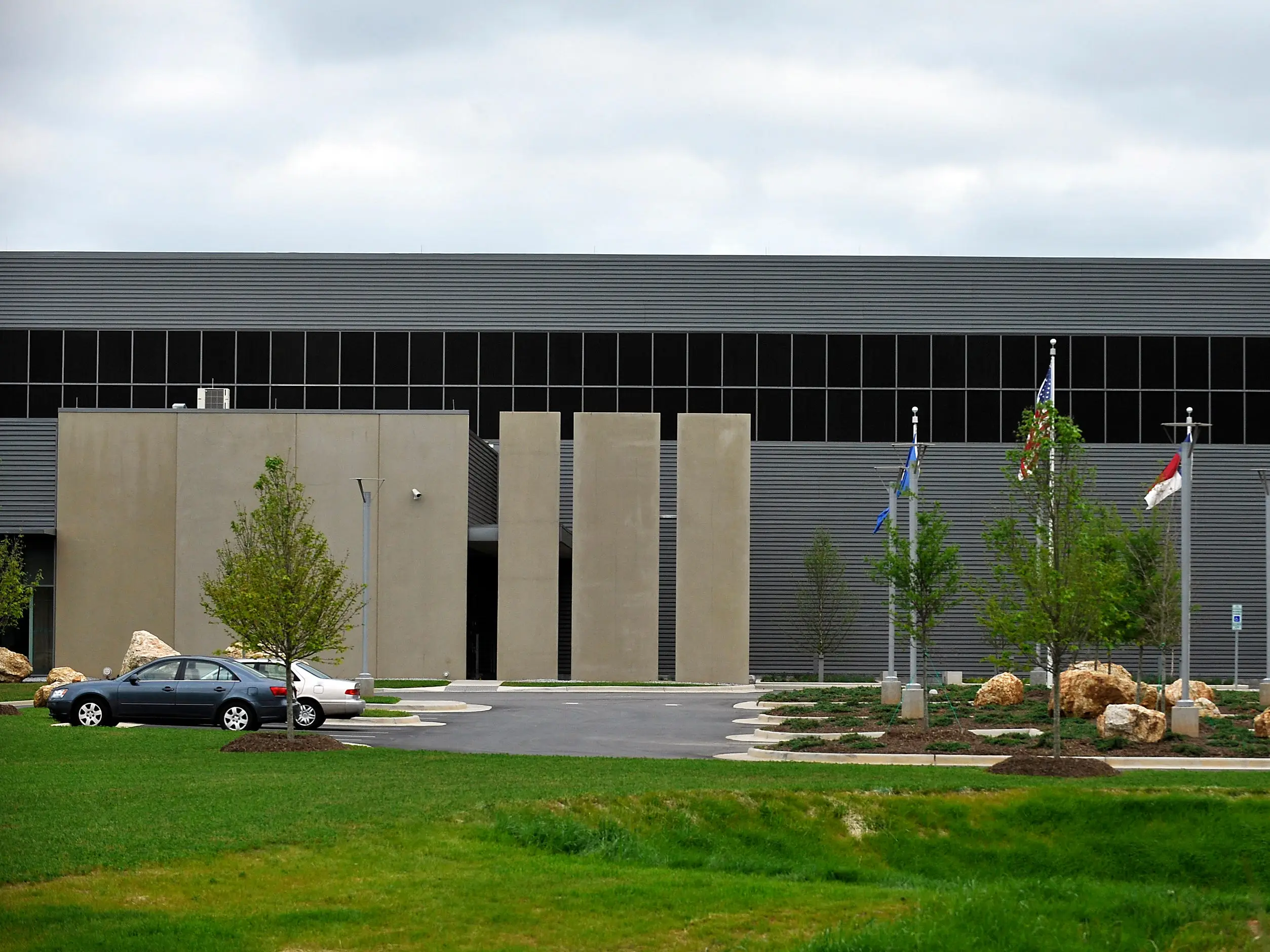 Modern gray building with vertical concrete panels, flags, parked cars, landscaped rocks, trees, and lawn under cloudy sky.