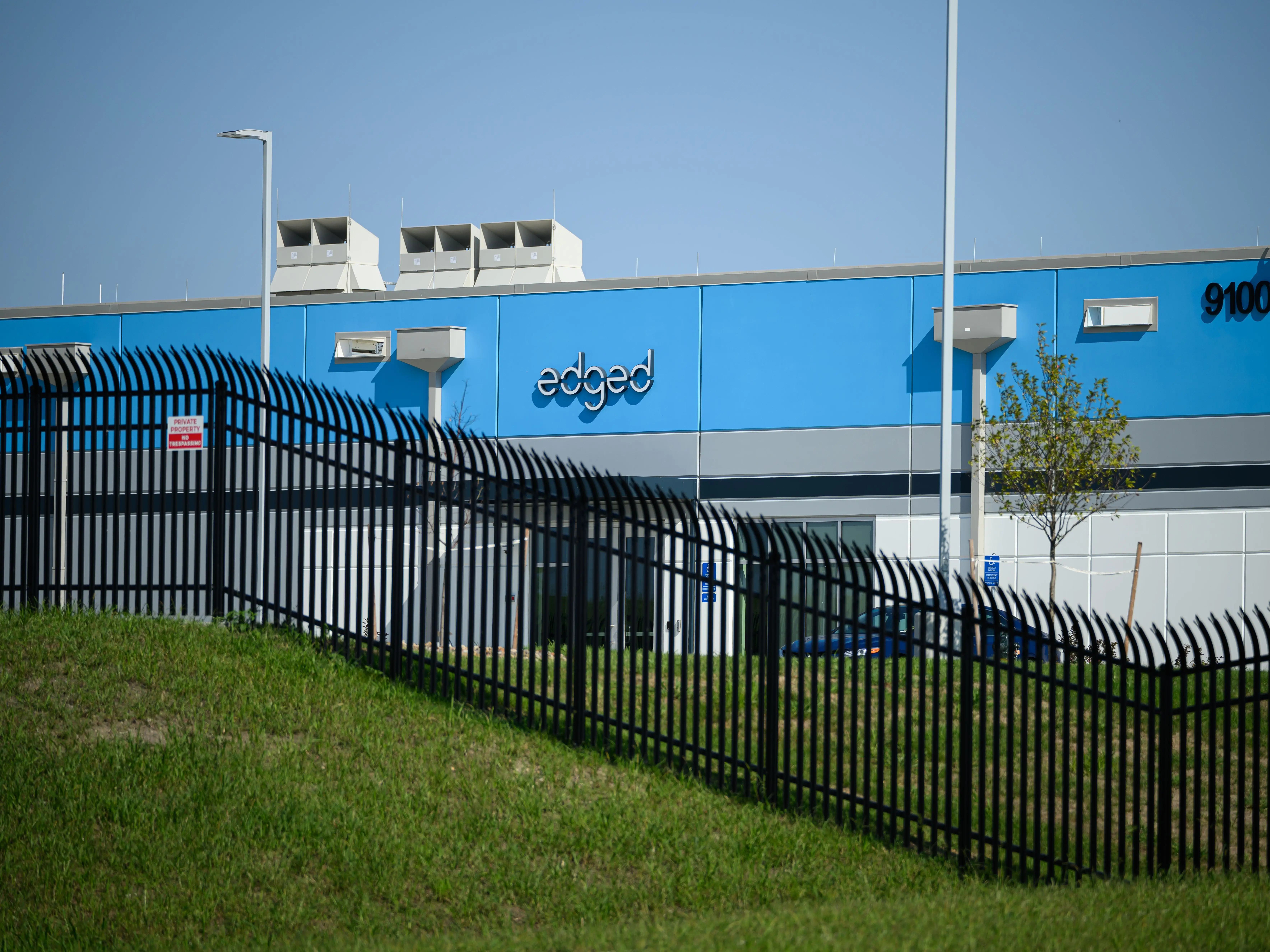 Blue and gray edged facility building at 9100 behind a black security fence under clear sky.