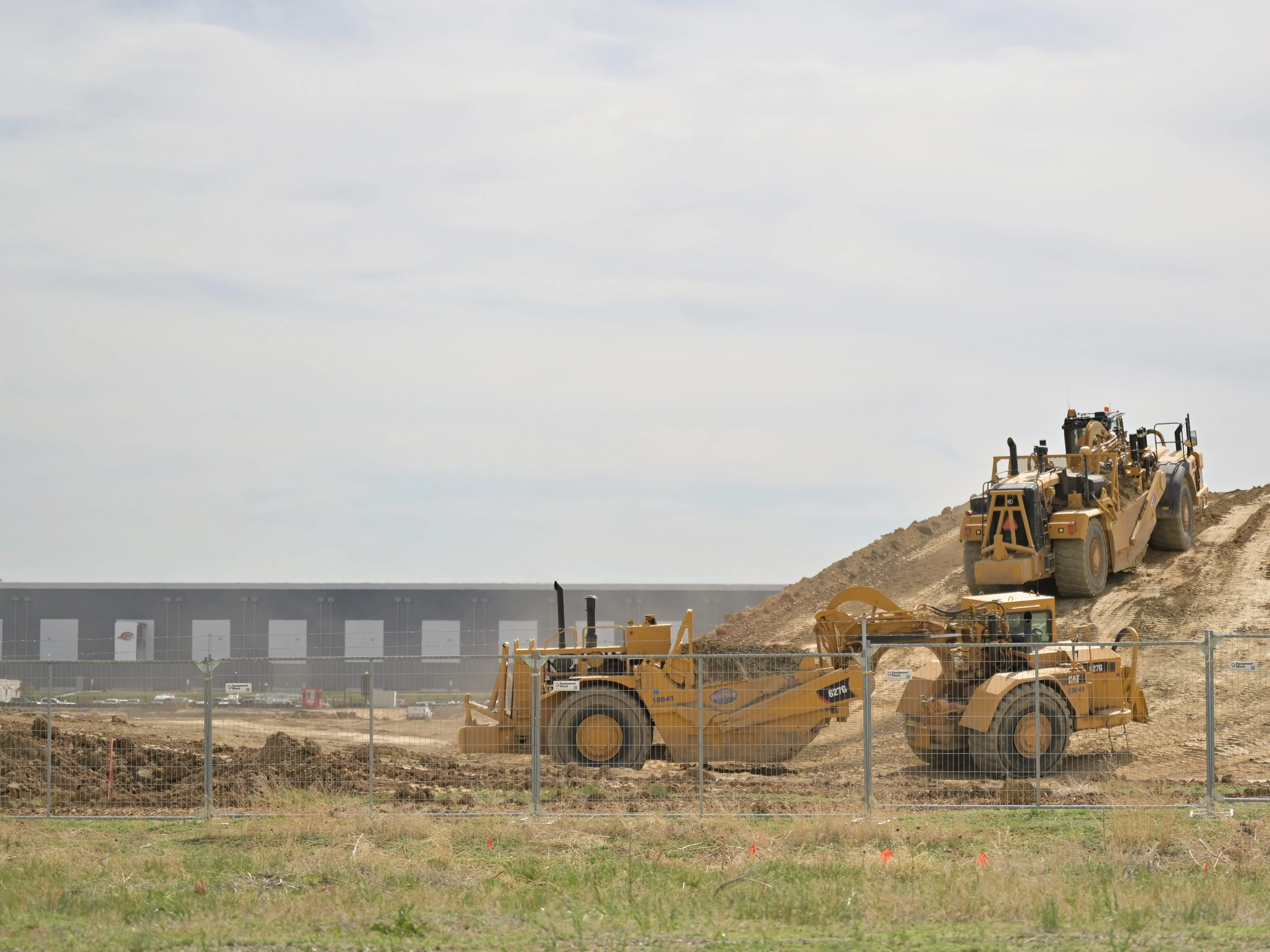 Heavy earthmoving machines work on a dirt mound behind a chain-link fence at a construction site.