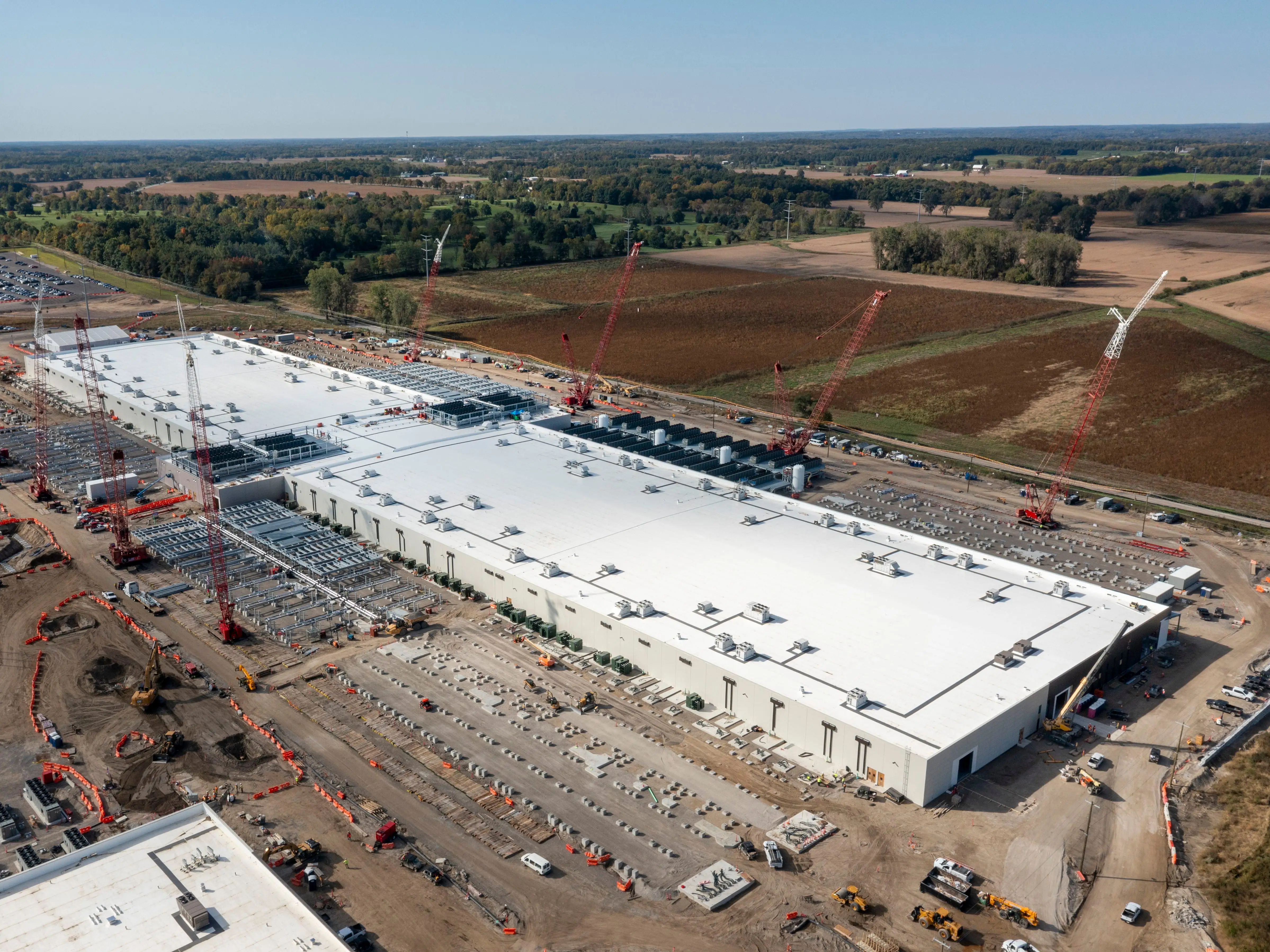 Aerial view of a large industrial construction site with cranes, white-roofed buildings, and surrounding farmland.