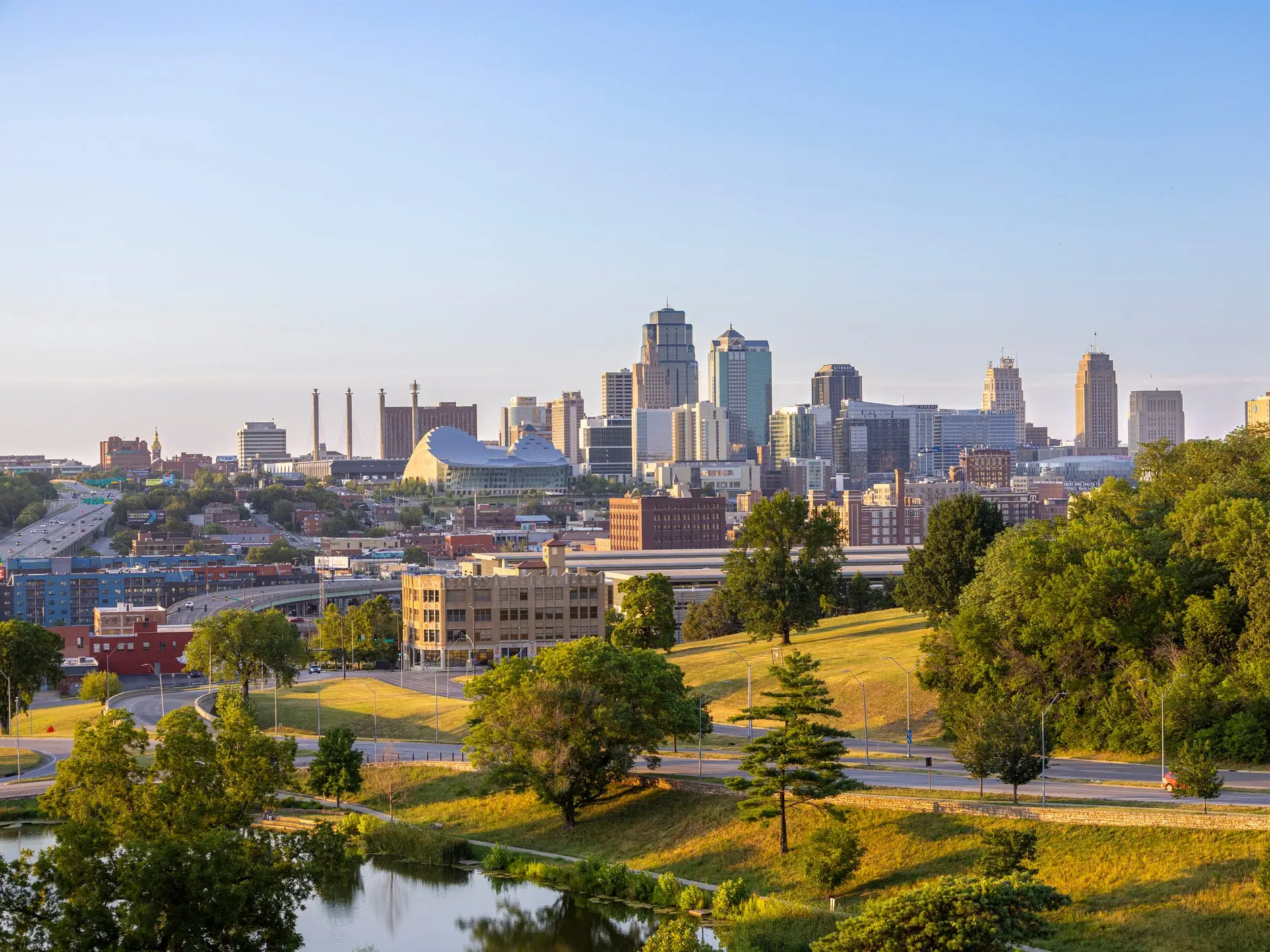 Downtown Kansas City skyline rises beyond green parkland and roads in warm evening light.