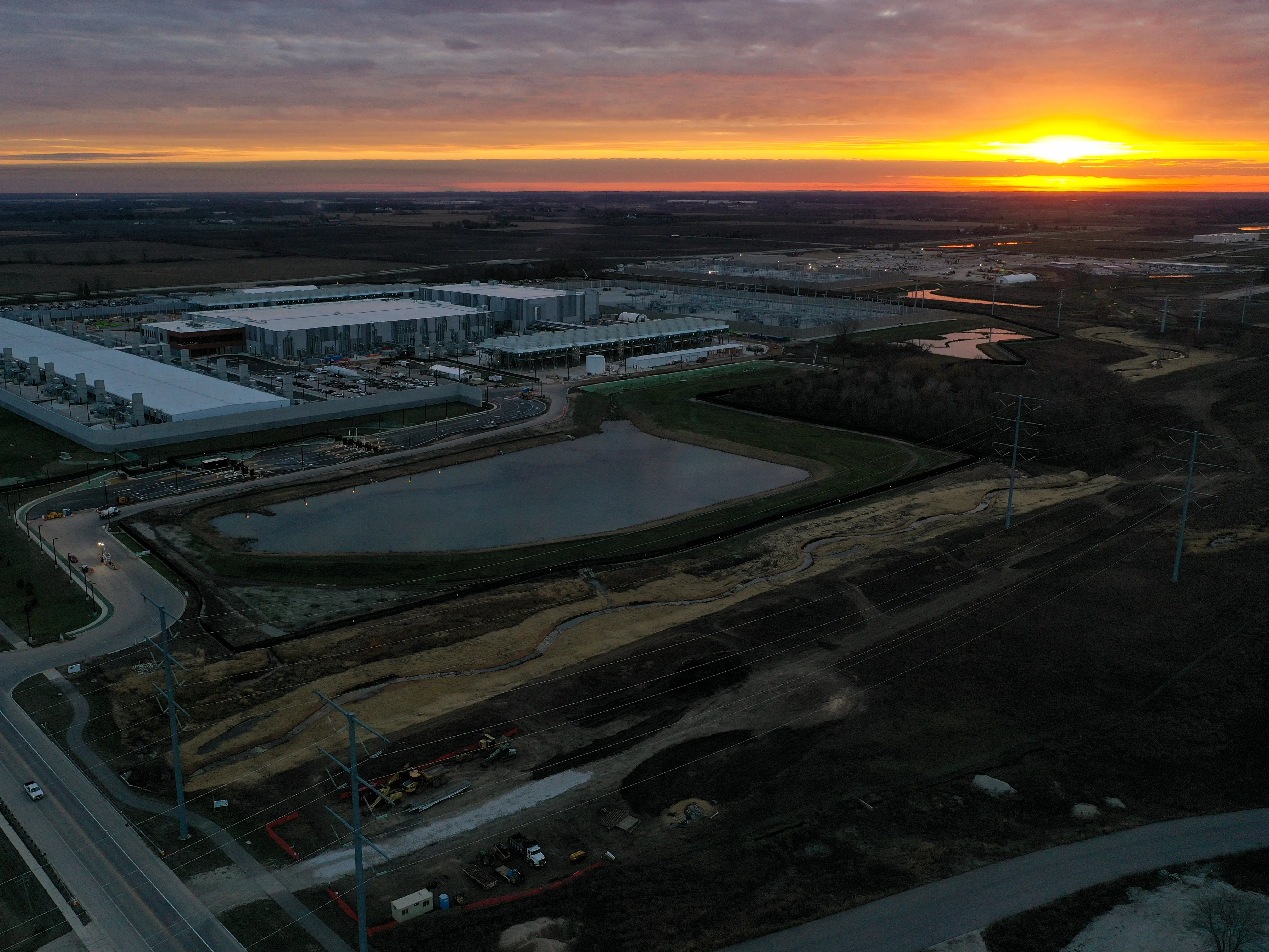Aerial view of a large industrial campus under construction with retention ponds, roads, and a sunset horizon.
