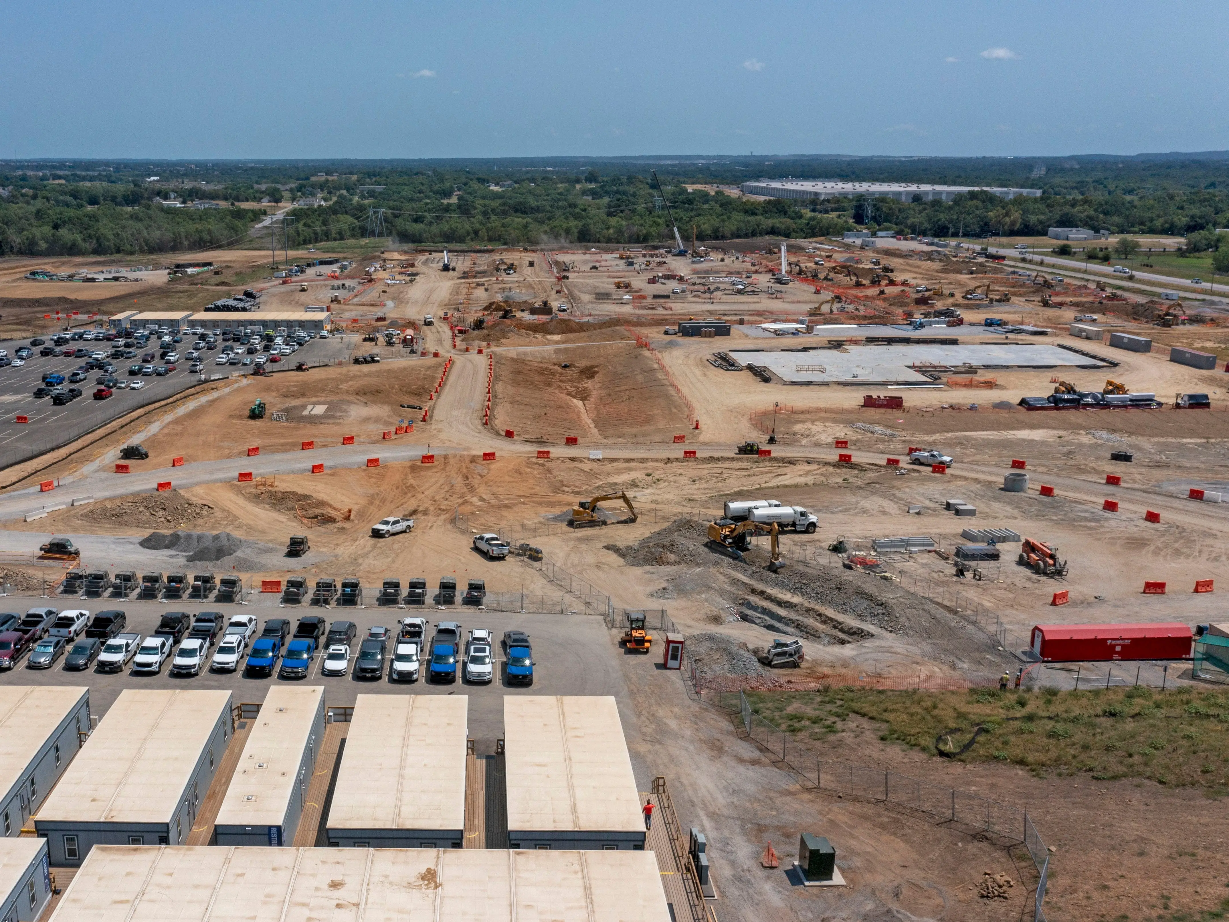 Aerial view of a large construction site with earthmoving equipment, parked cars, trailers, and cleared land.