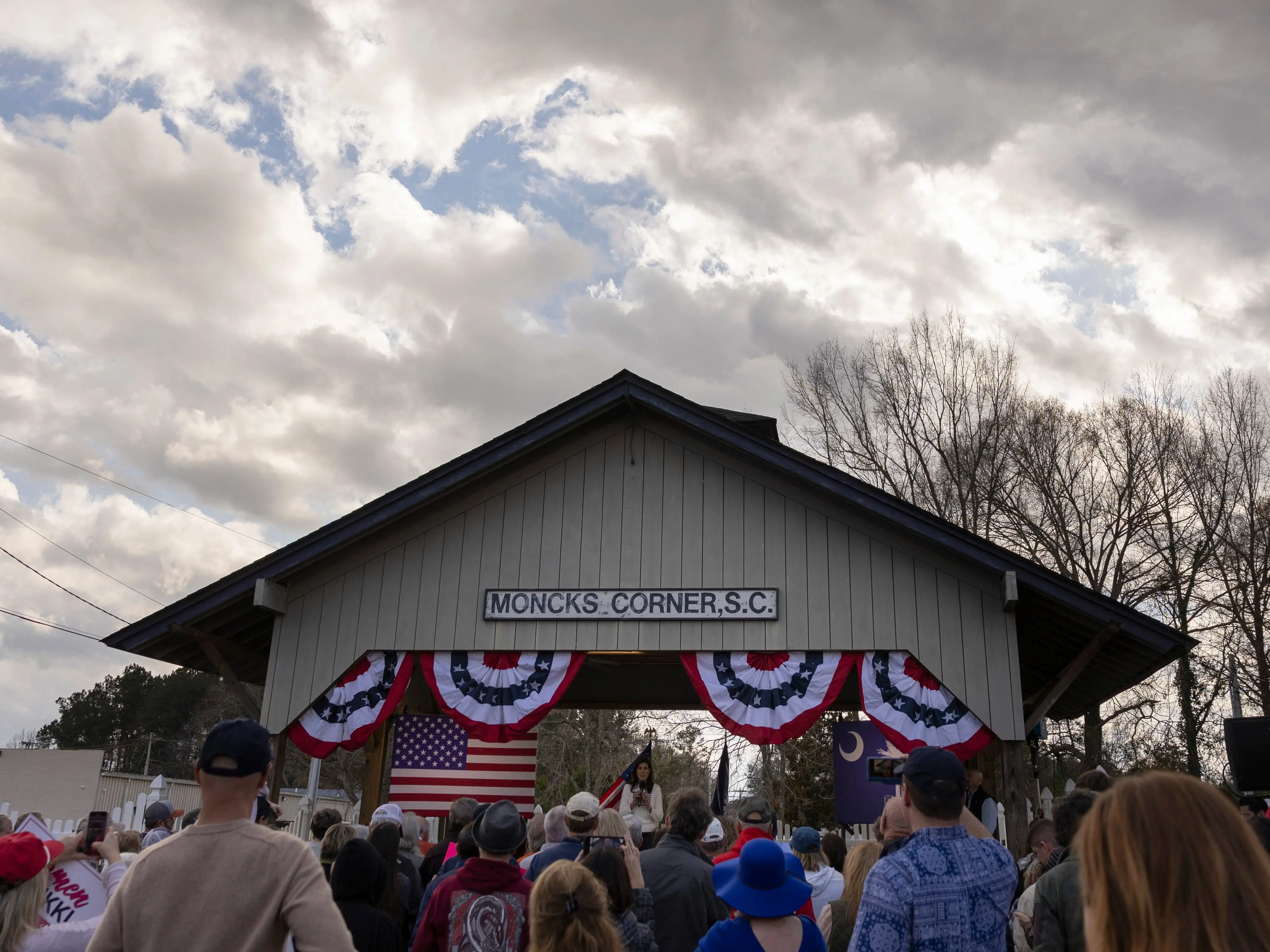 Crowd gathers before a bunting-decorated stage labeled “MONCKS CORNER,S.C.” under a cloudy sky.