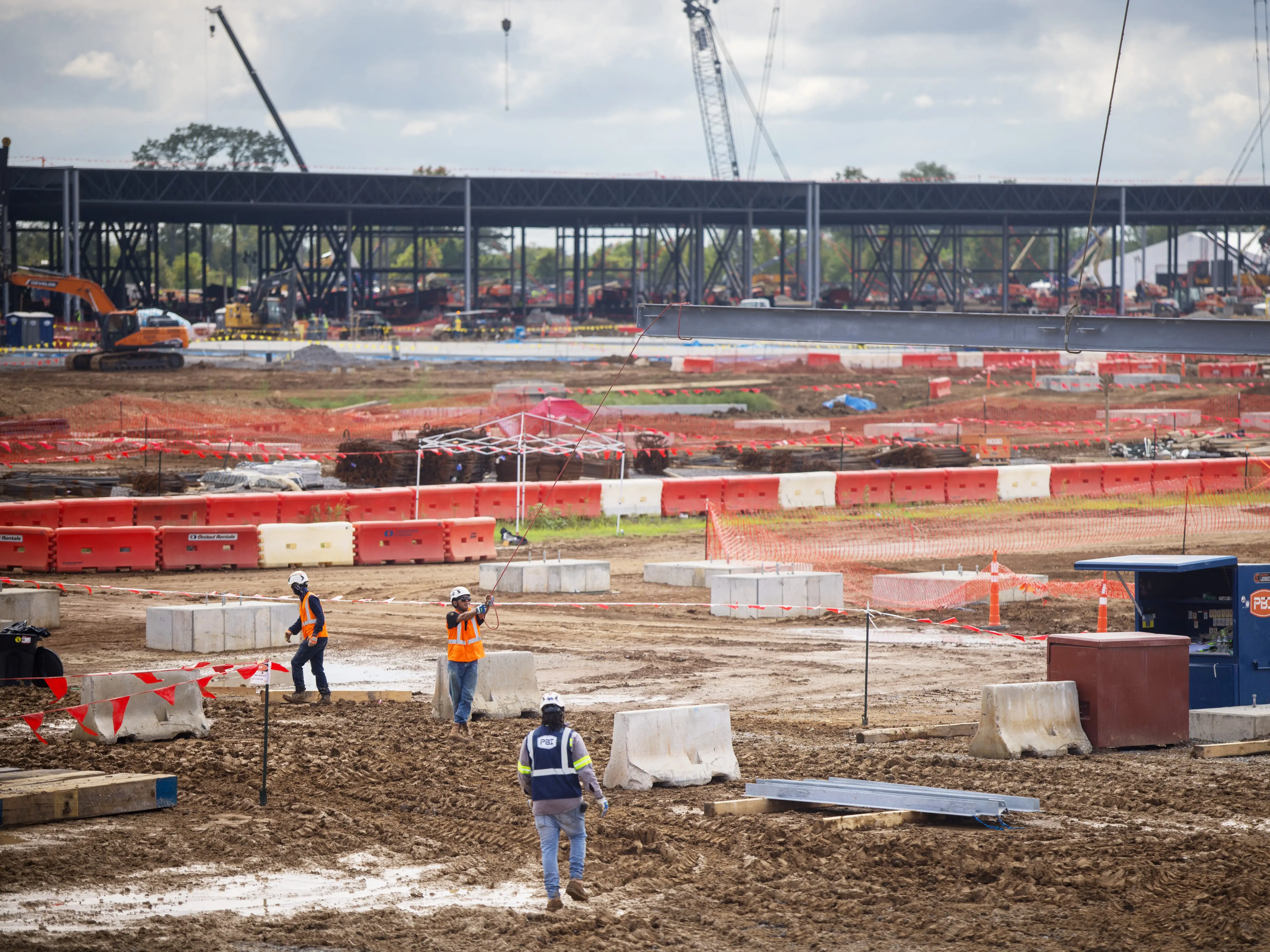 Workers in safety vests walk through a muddy construction site with cranes, barriers, and steel framing in the background.