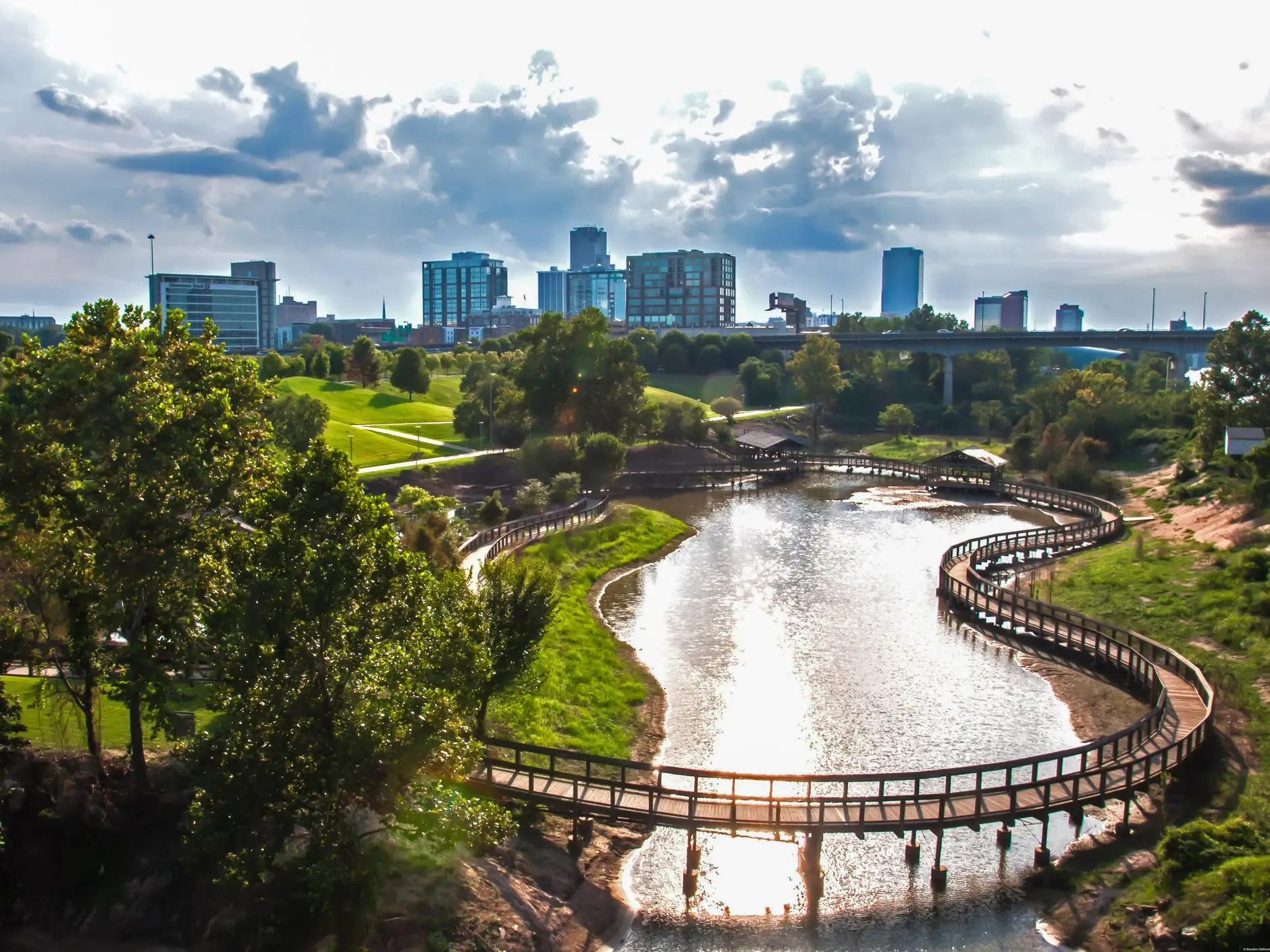 Urban park with a winding boardwalk over water, green trees, and a modern skyline beneath cloudy sunlight.