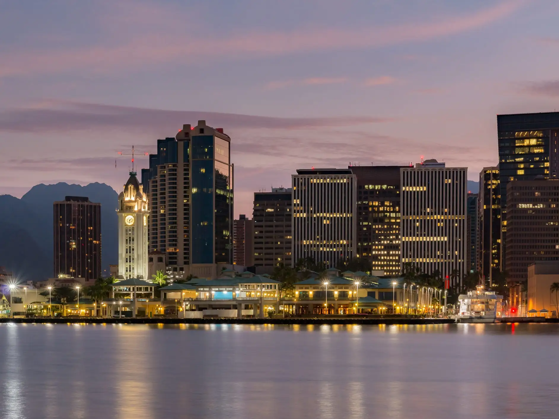Honolulu waterfront skyline at dusk with Aloha Tower lit beside harbor water and office buildings.