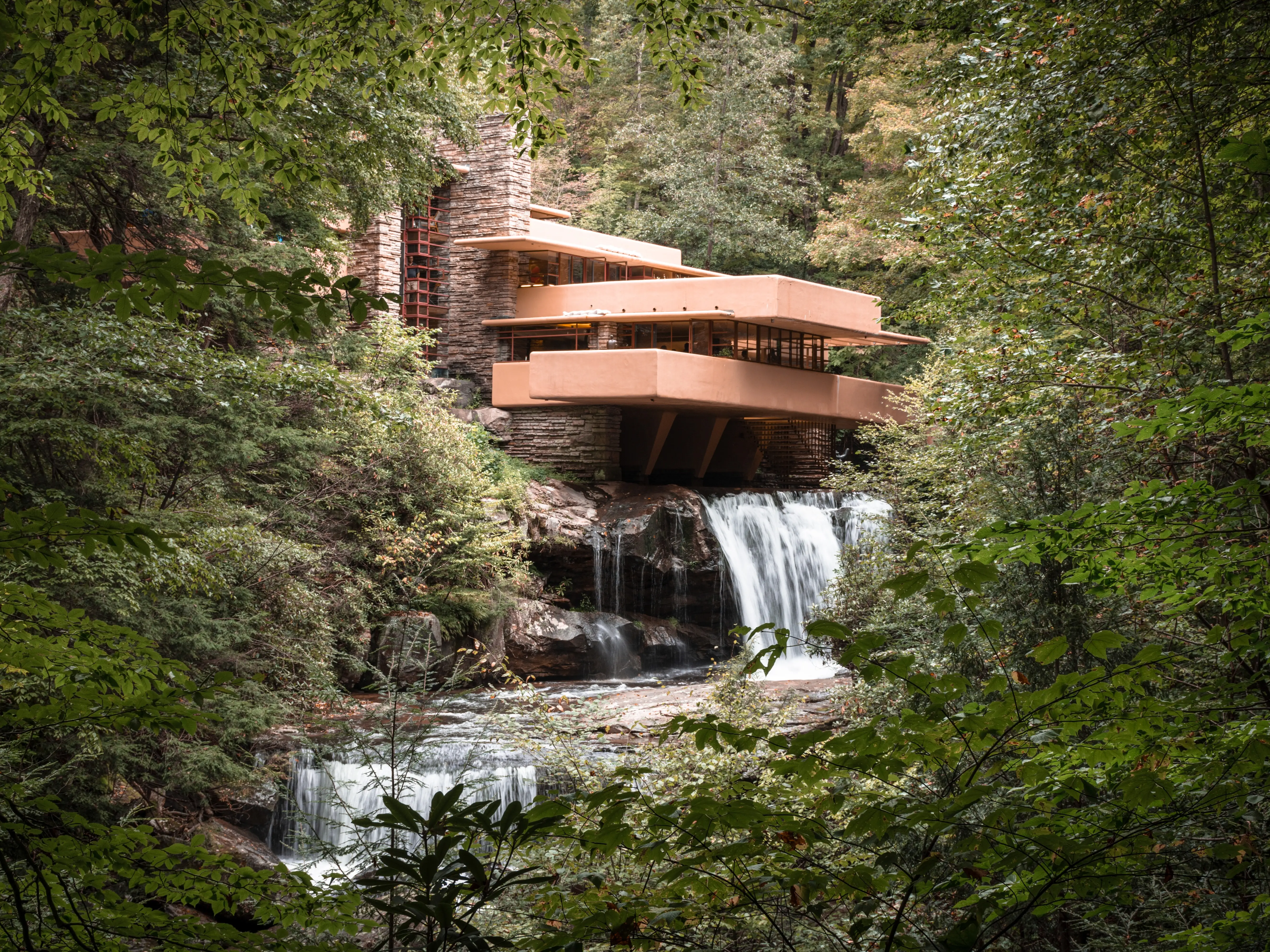 Frank Lloyd Wright - Fallingwater, view from the woods and stream from below towards the house with lush foliage and two waterfalls