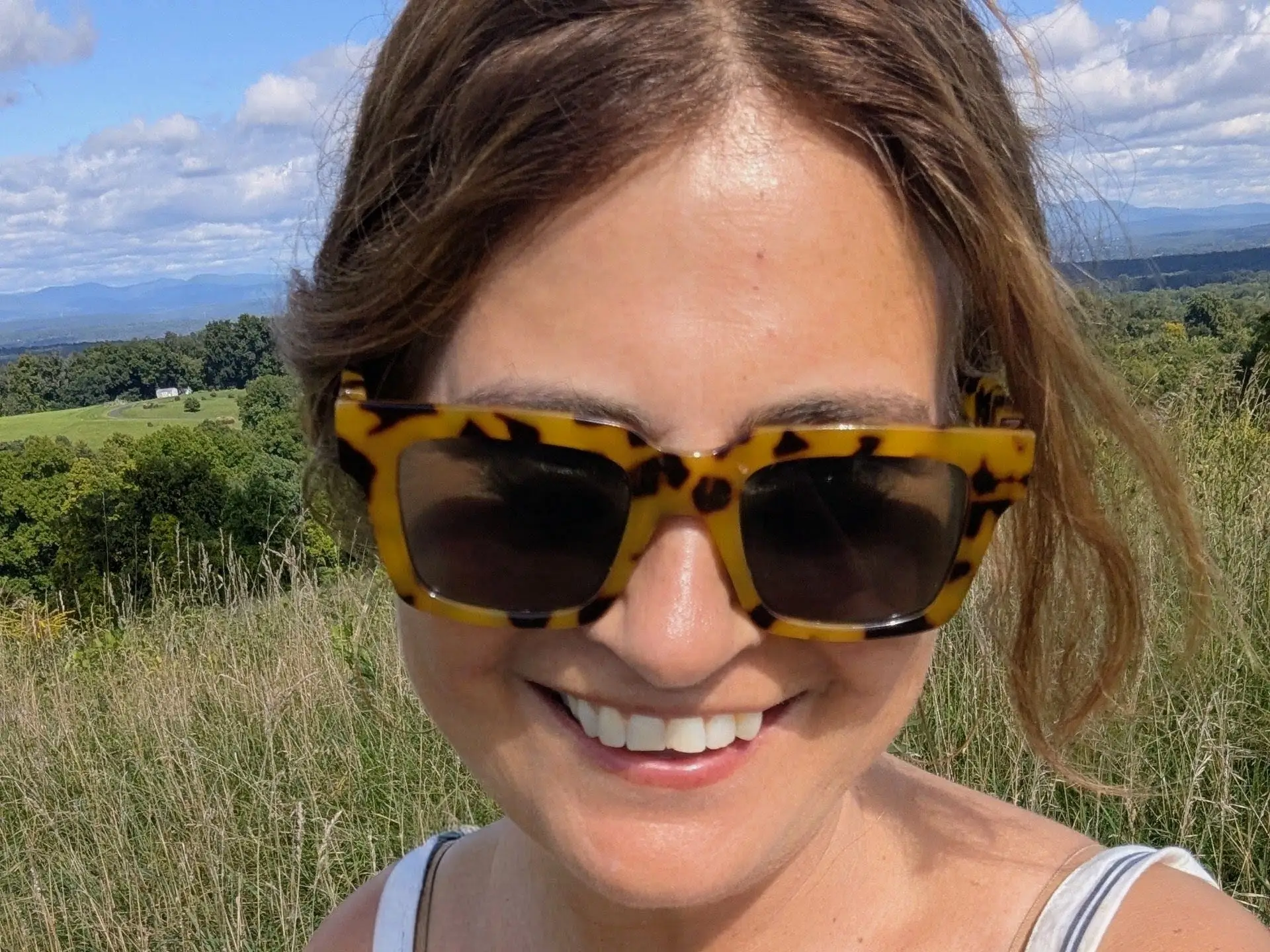 Smiling person wearing tortoiseshell sunglasses stands in a grassy field with rolling countryside under a partly cloudy sky.