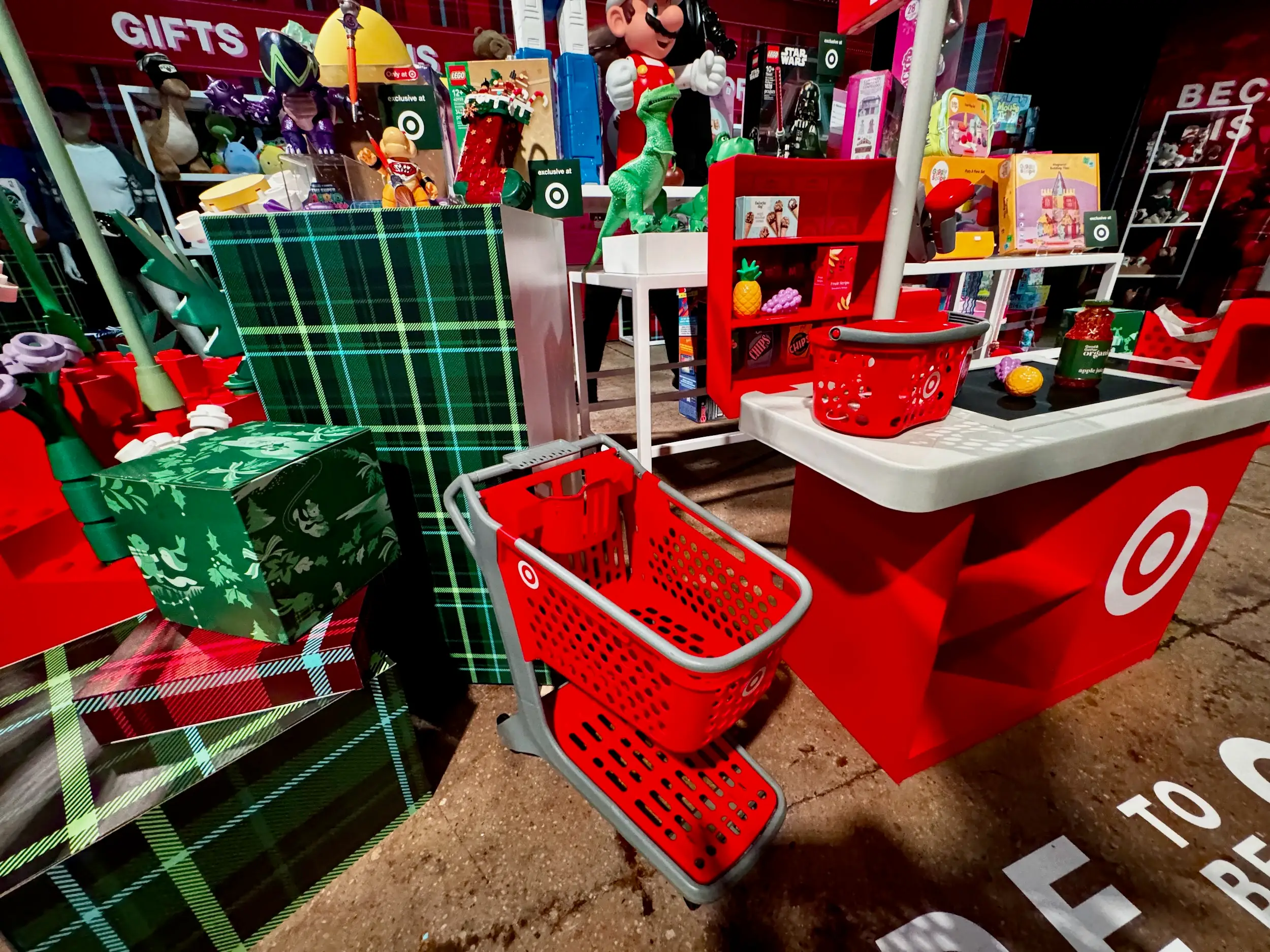 Target holiday gift display with red shopping carts, wrapped presents, toys, and checkout counter under bright store lighting.