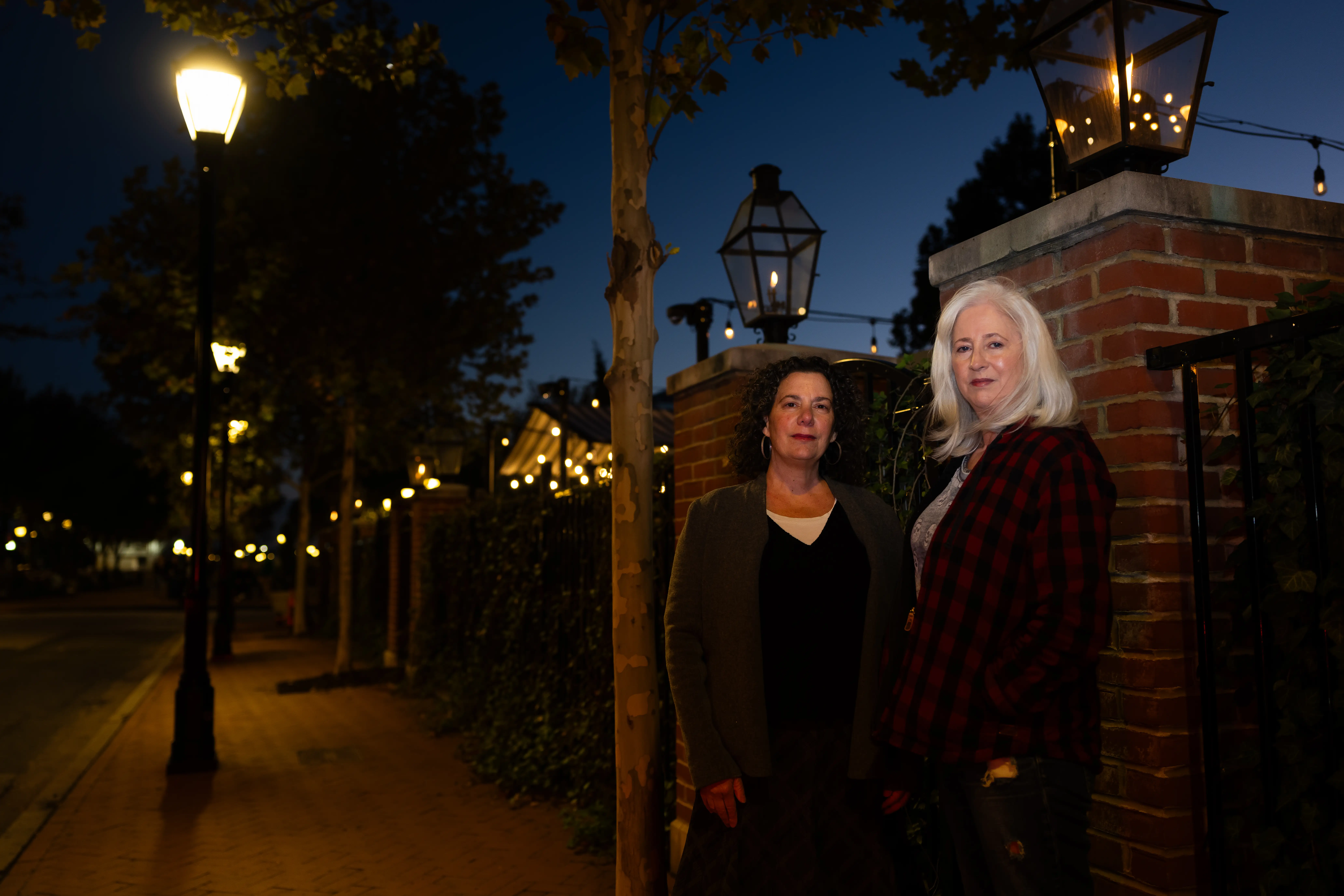 Two people stand beside a brick wall on a lamplit sidewalk at dusk.