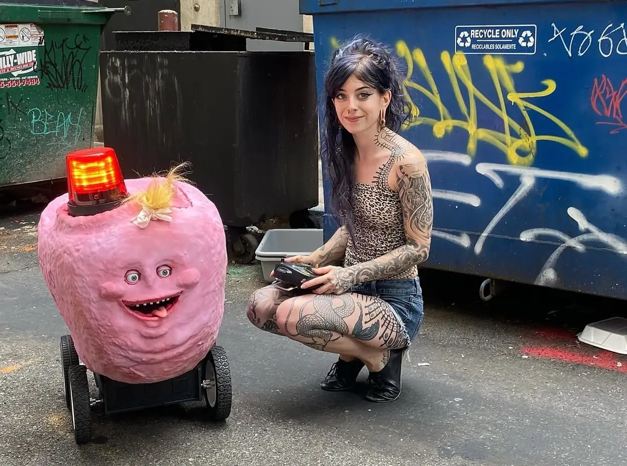 A tattooed person crouches beside a pink fuzzy robot with an orange beacon in a graffiti-marked alley.