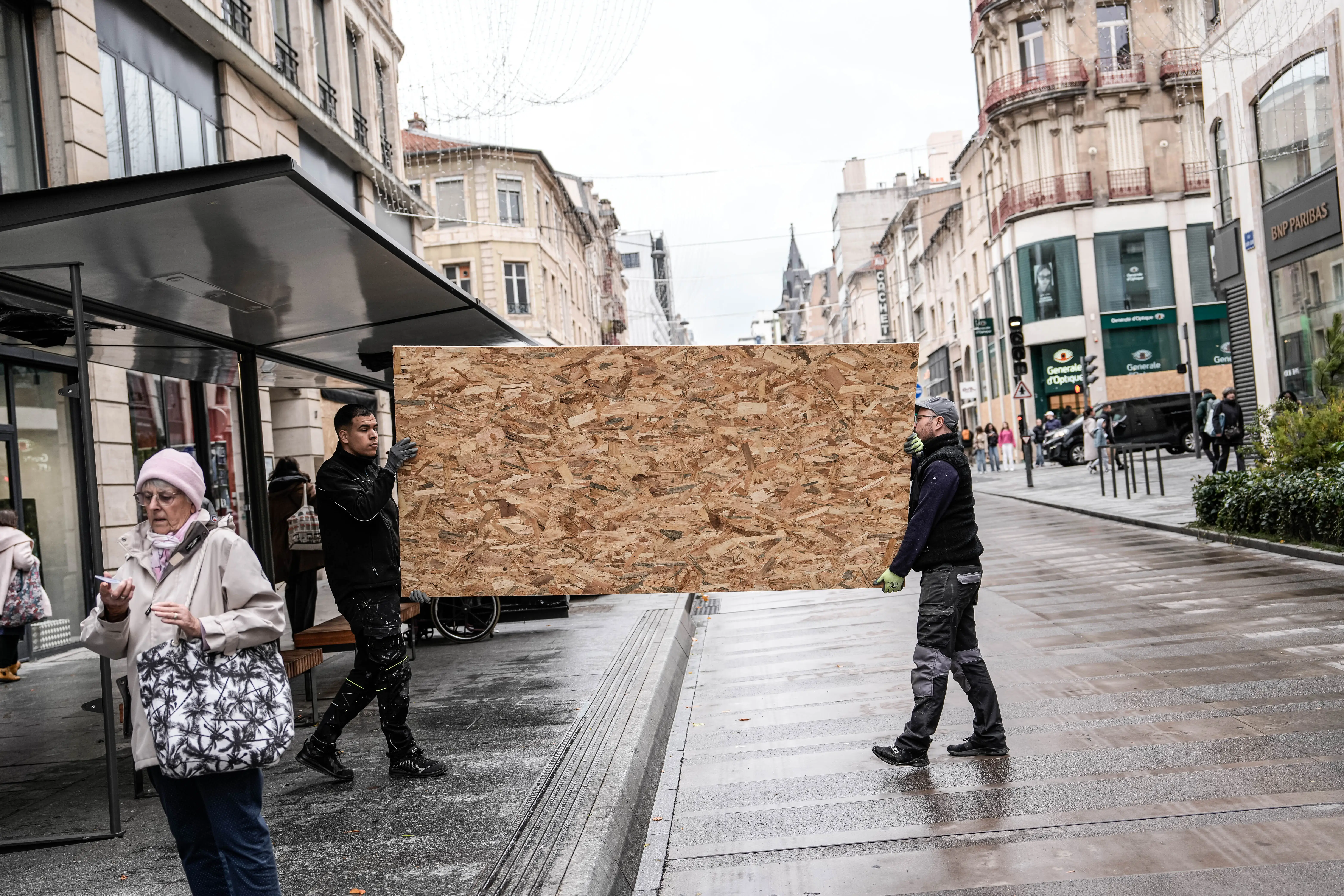 Two men carry a large piece of plywood down a French street