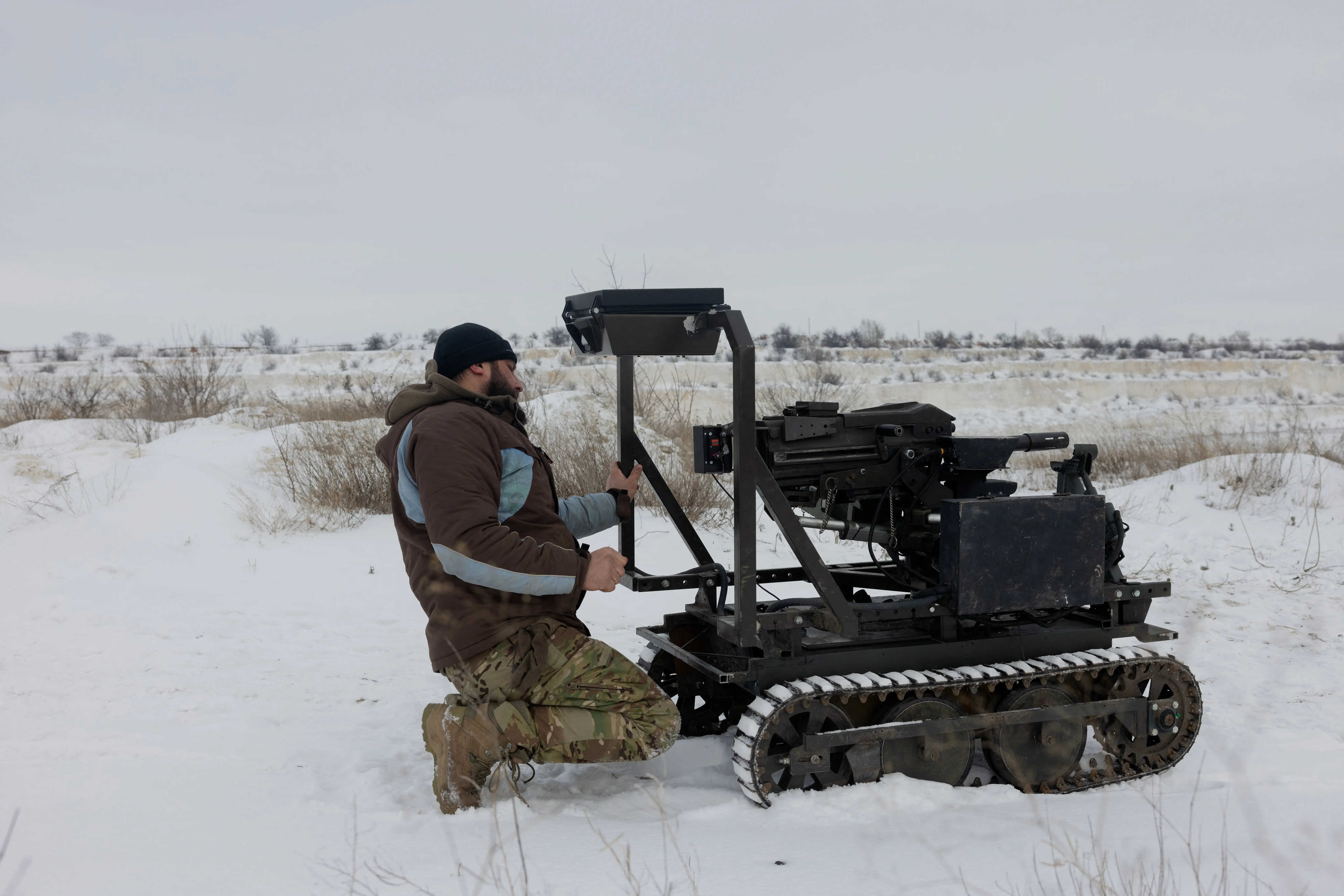 A person kneels beside a tracked robotic vehicle in a snowy open field under an overcast sky.