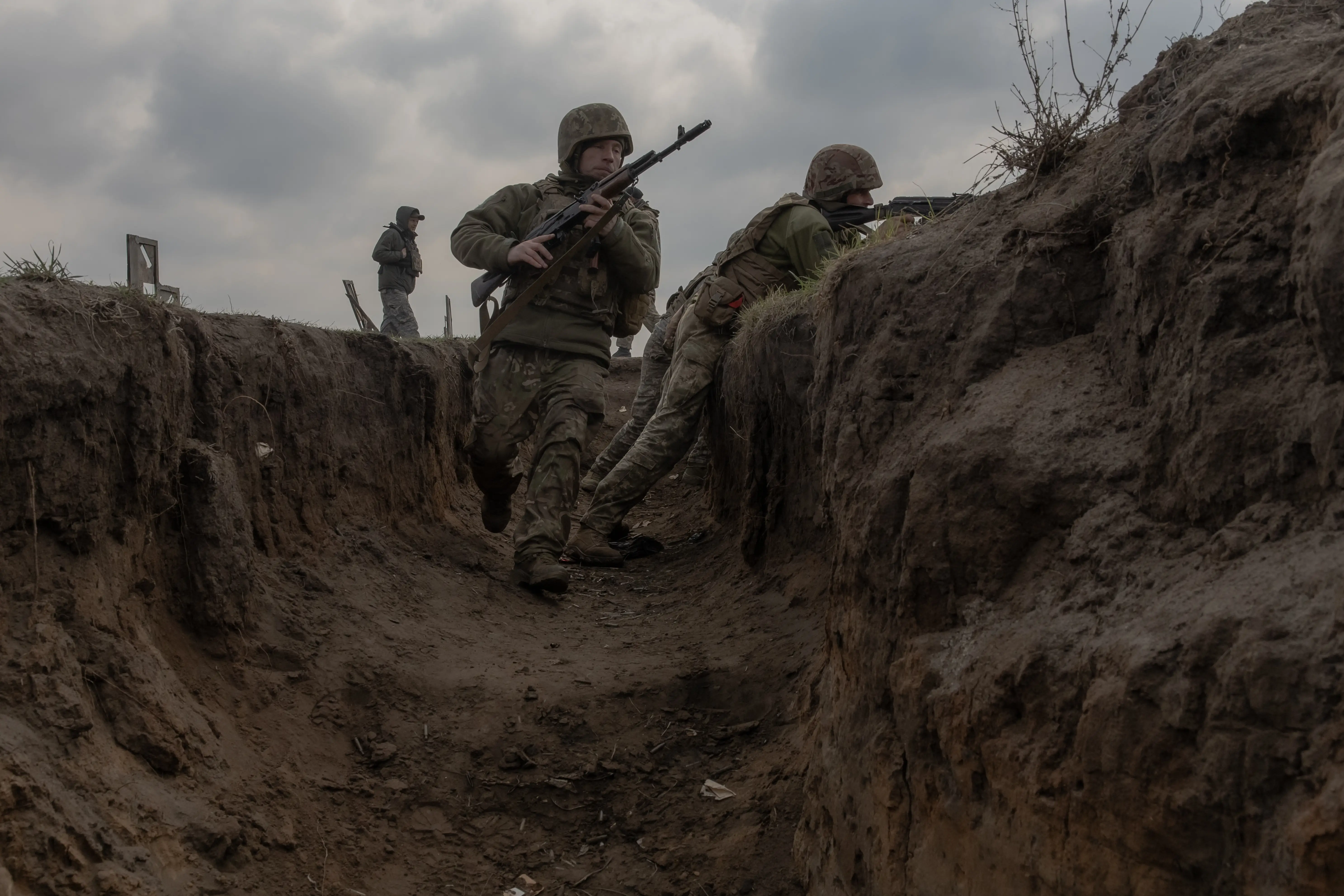Helmeted soldiers carrying rifles move through a muddy trench under a cloudy sky.