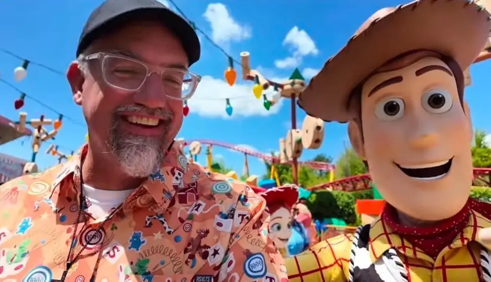 Smiling person poses beside Woody in a Toy Story themed outdoor amusement park area.