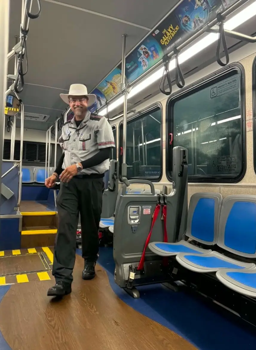 Uniformed transit employee walks through a mostly empty vehicle with blue seats, grab straps, and overhead movie advertising.