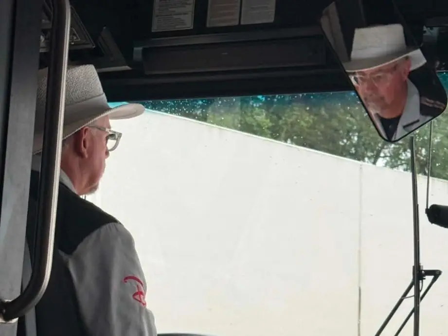 Uniformed transit operator sits at the controls inside a bus with windshield, mirror, and dashboard displays visible.