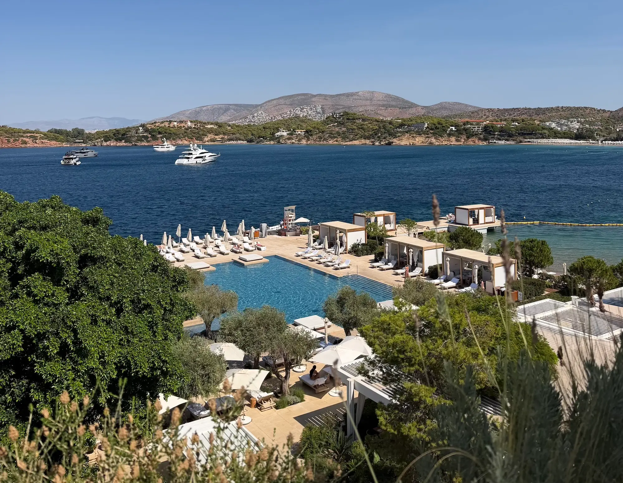 Seaside resort pool and cabanas overlook a blue bay with yachts and hills in the distance.