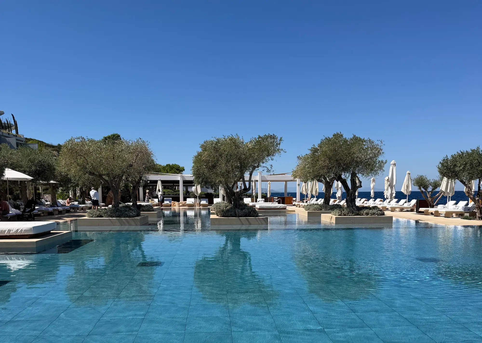 Resort swimming pool with sun loungers, closed umbrellas, trees, and sea views under a clear blue sky.