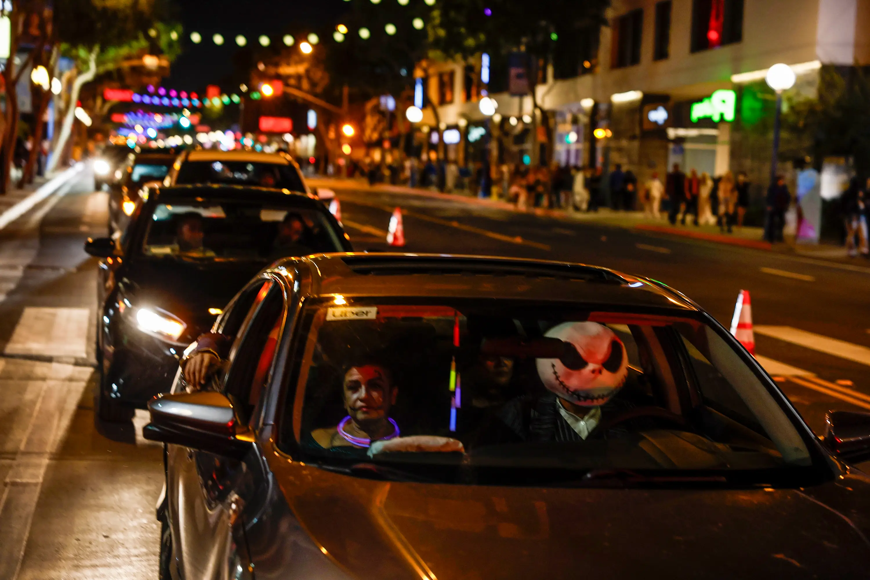 Cars line a brightly lit city street at night with passengers visible through a windshield.