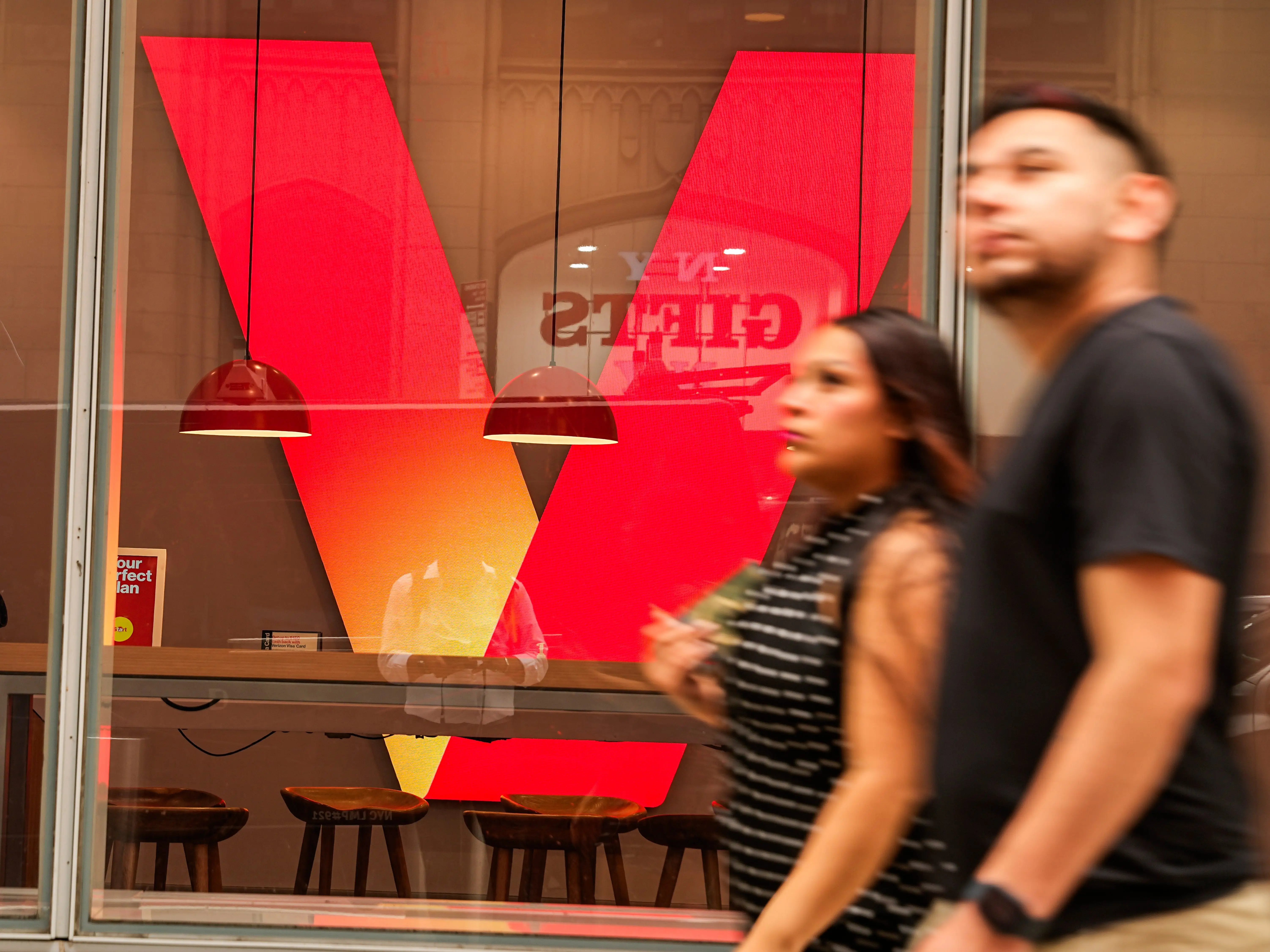 Pedestrians pass a Verizon storefront window with a large red logo and a digital display showing a red robot.