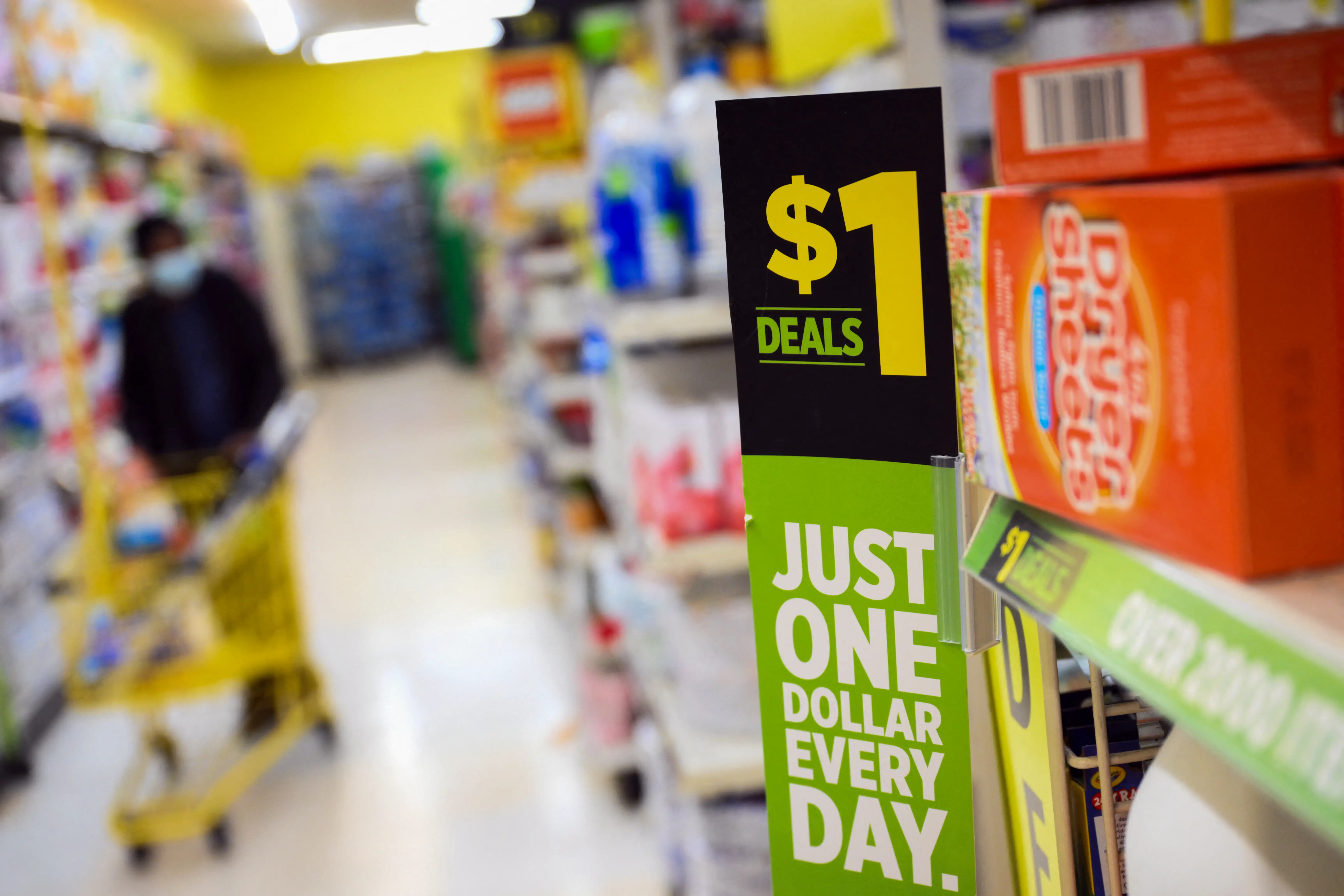 Dollar store aisle shelf sign advertises $1 deals beside orange packaged goods as a shopper pushes a cart nearby.