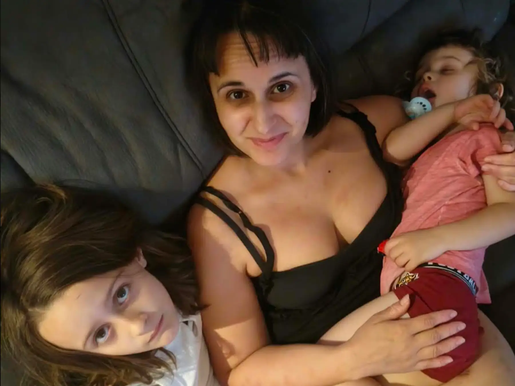 Three people lie together on a gray couch, with one resting while using a pacifier.