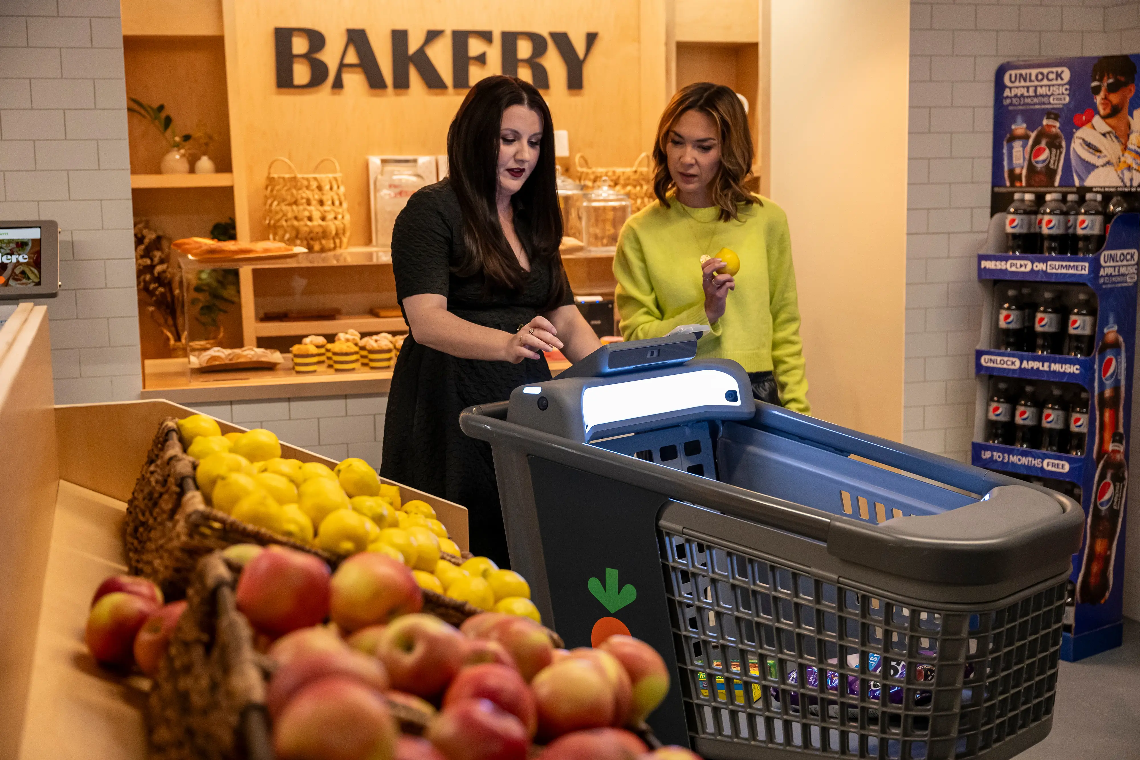 Two shoppers use a screen-equipped shopping cart beside apples and lemons near a bakery section.