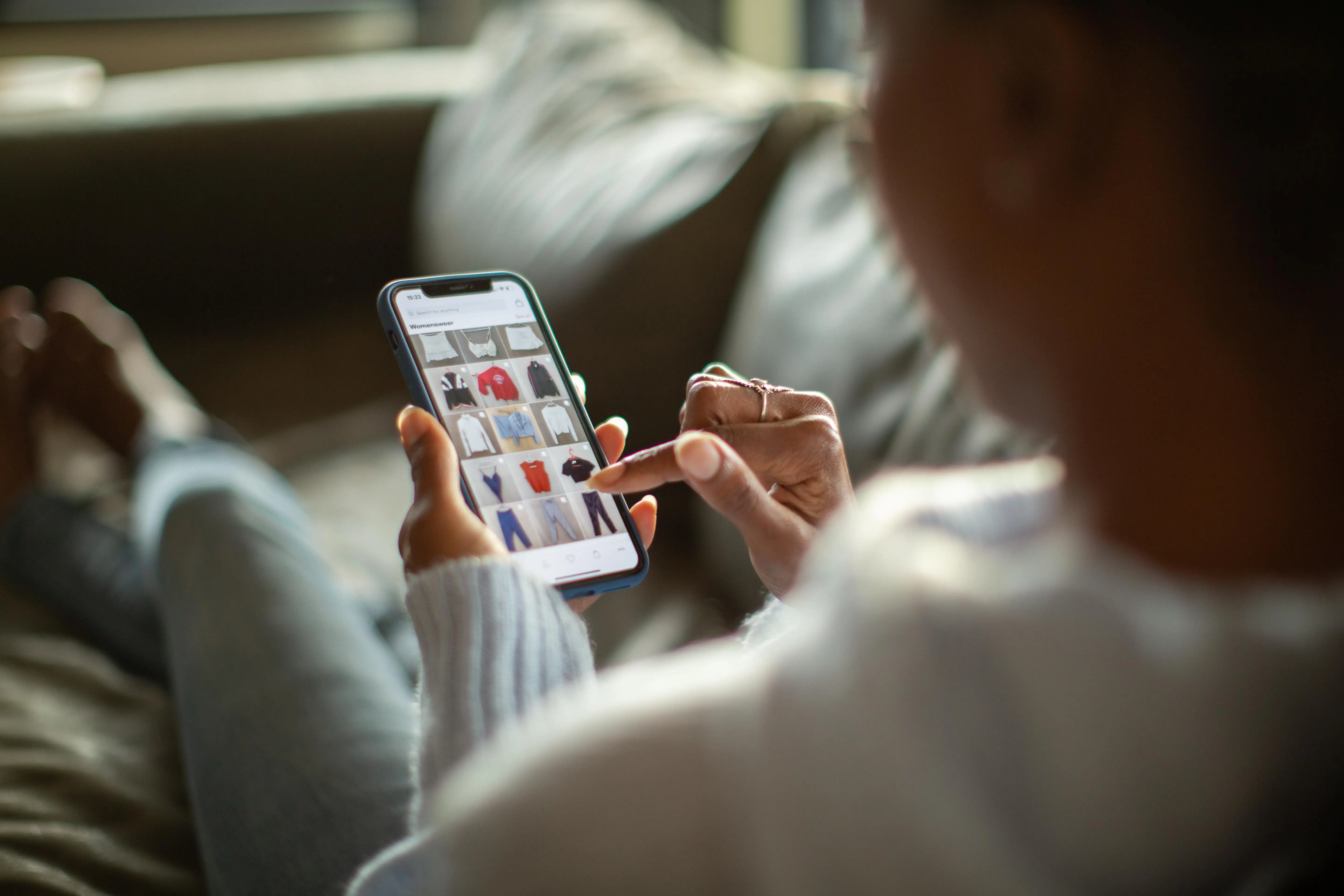 Person seated on a couch browsing clothing items on a smartphone shopping interface.