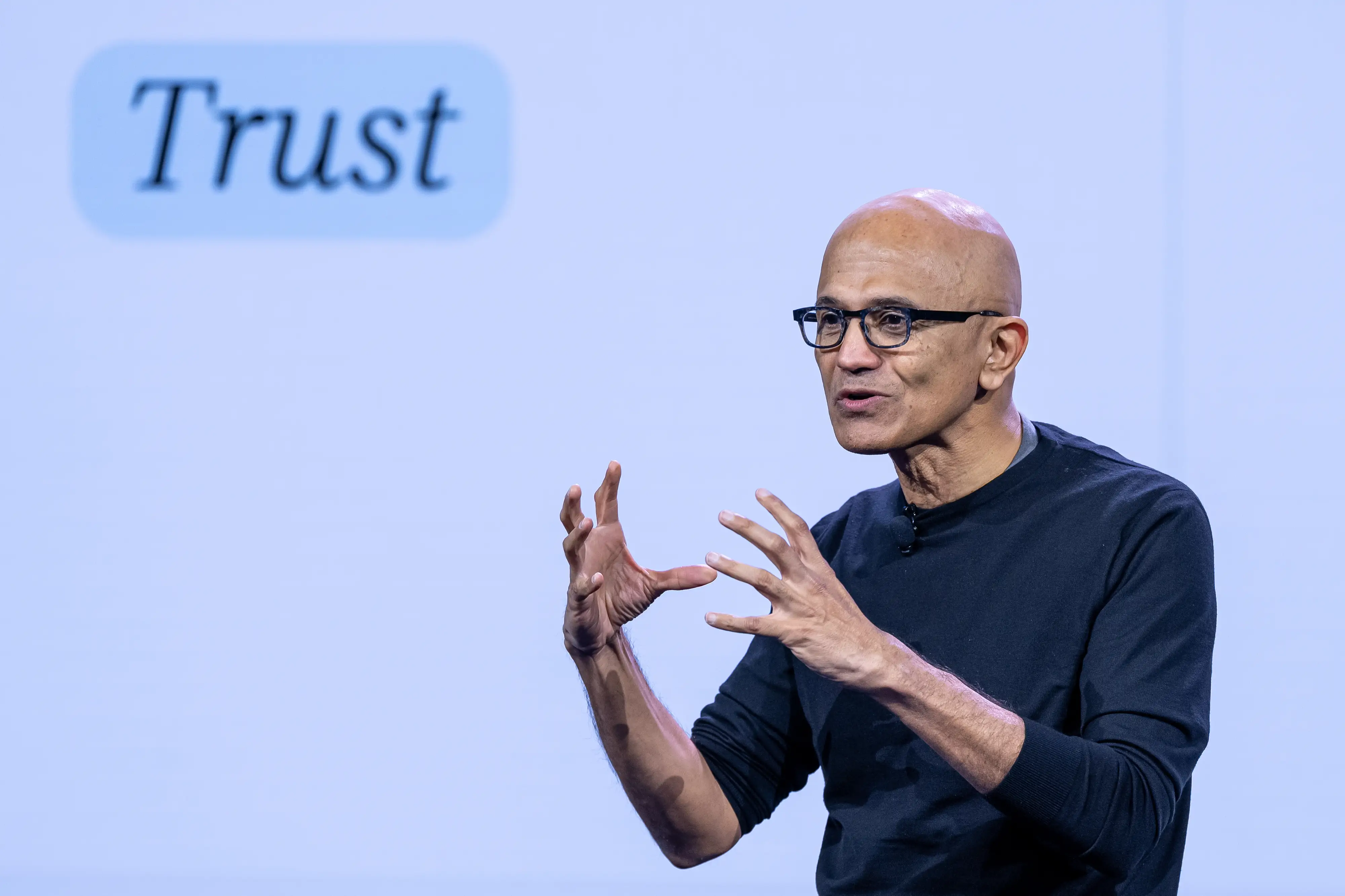 Satya Nadella speaks onstage beside a projected slide reading “Trust.”
