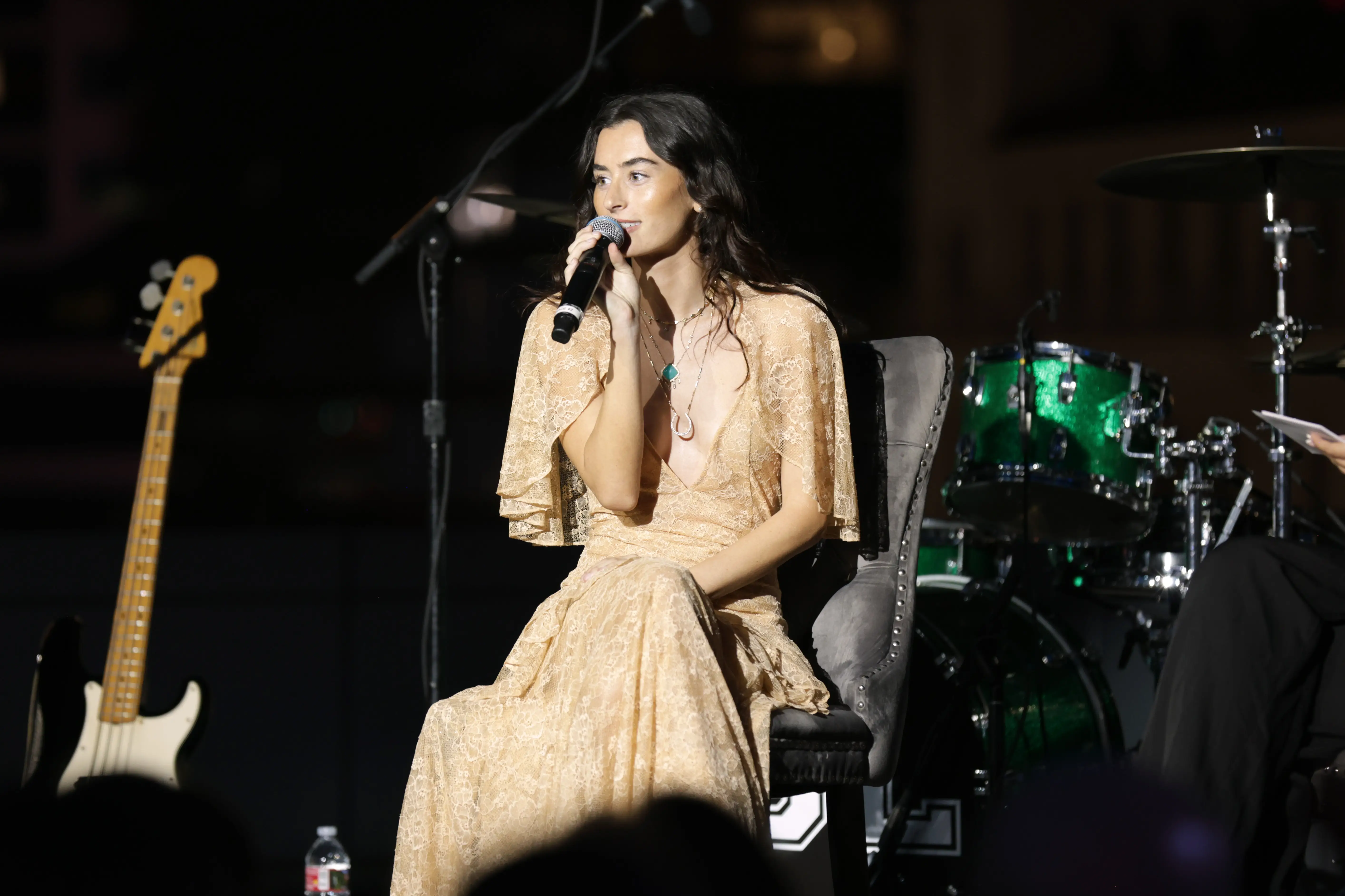 Stella Lefty sits onstage singing into a handheld microphone with drums and a bass guitar nearby.