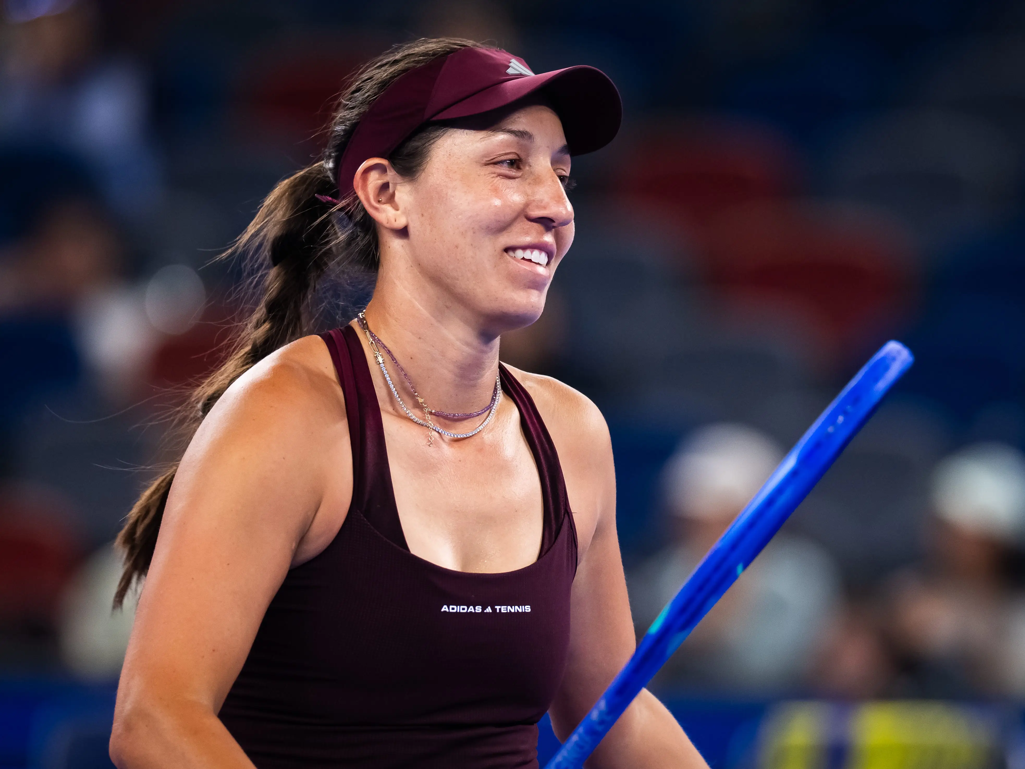 Jessica Pegula smiles on a tennis court while holding a blue training stick during a match or practice setting.