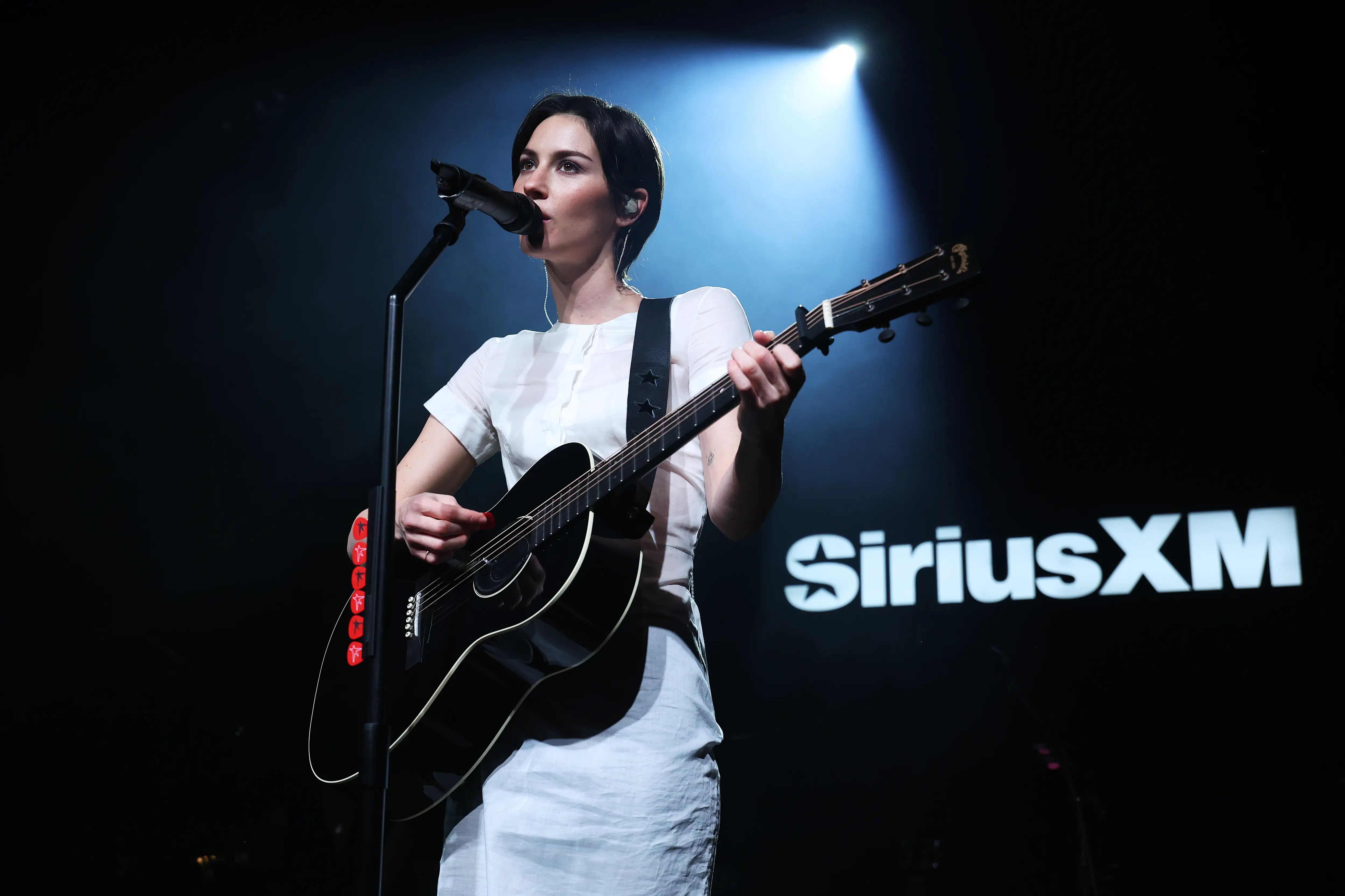 St. Vincent performs with a black acoustic guitar on a dark stage with a SiriusXM backdrop.