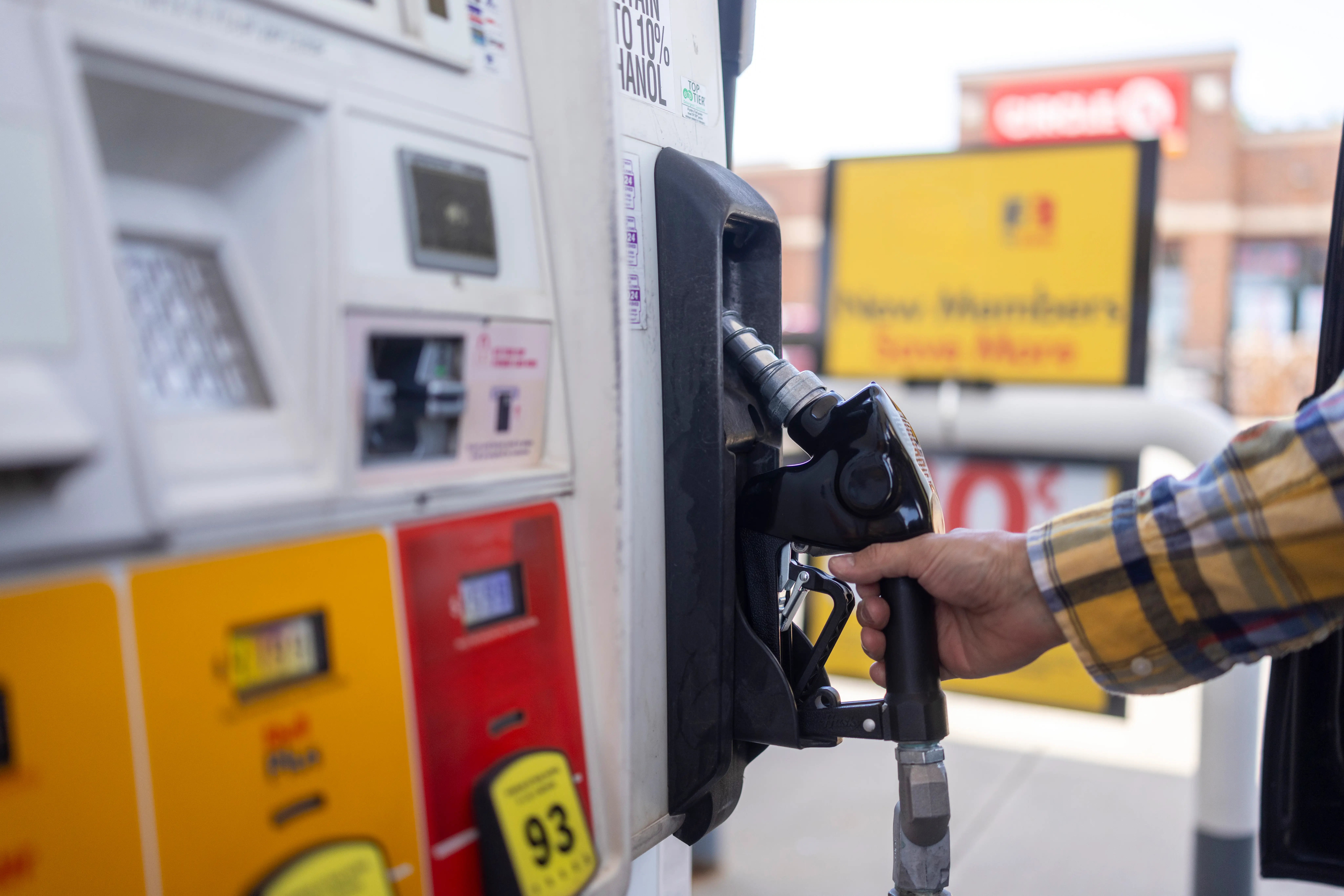 A hand in a plaid shirt holding a fuel nozzle refueling a car at a gas station pump. Bright day with a visible station backdrop.