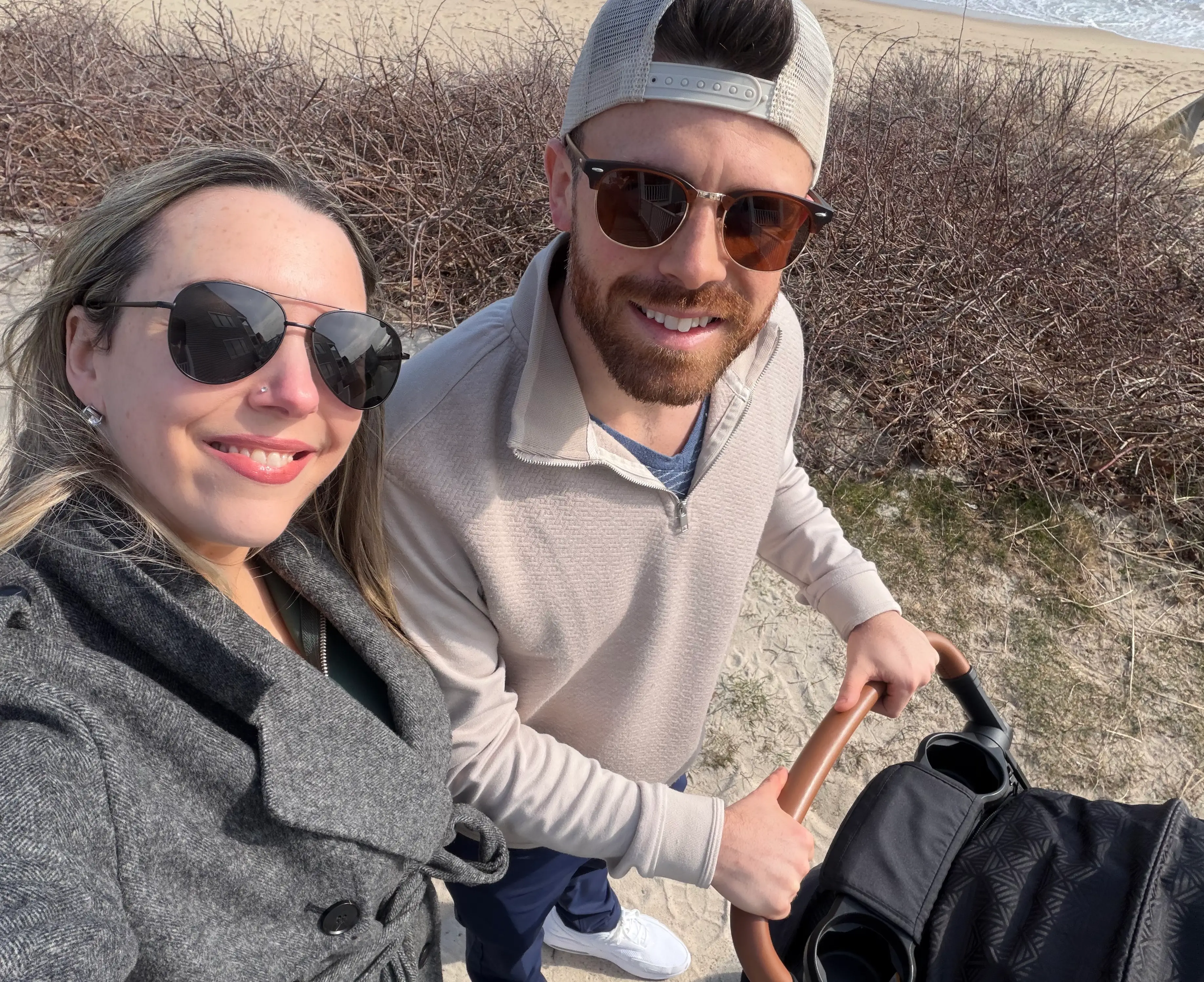 Two adults wearing sunglasses smile for a beachside selfie while pushing a stroller on a sandy path.