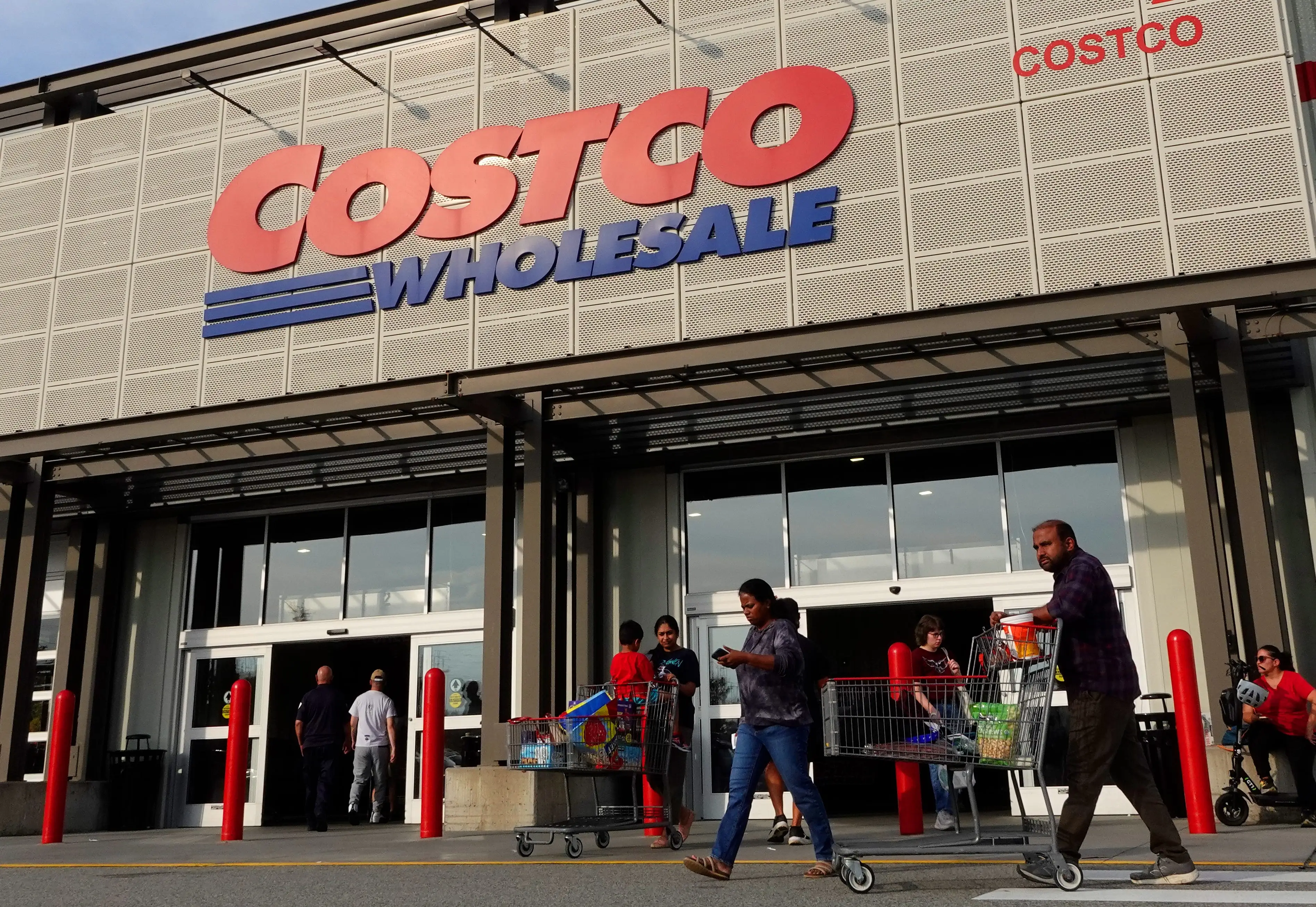 Shoppers push carts outside a Costco Wholesale warehouse entrance with large red and blue storefront signage.