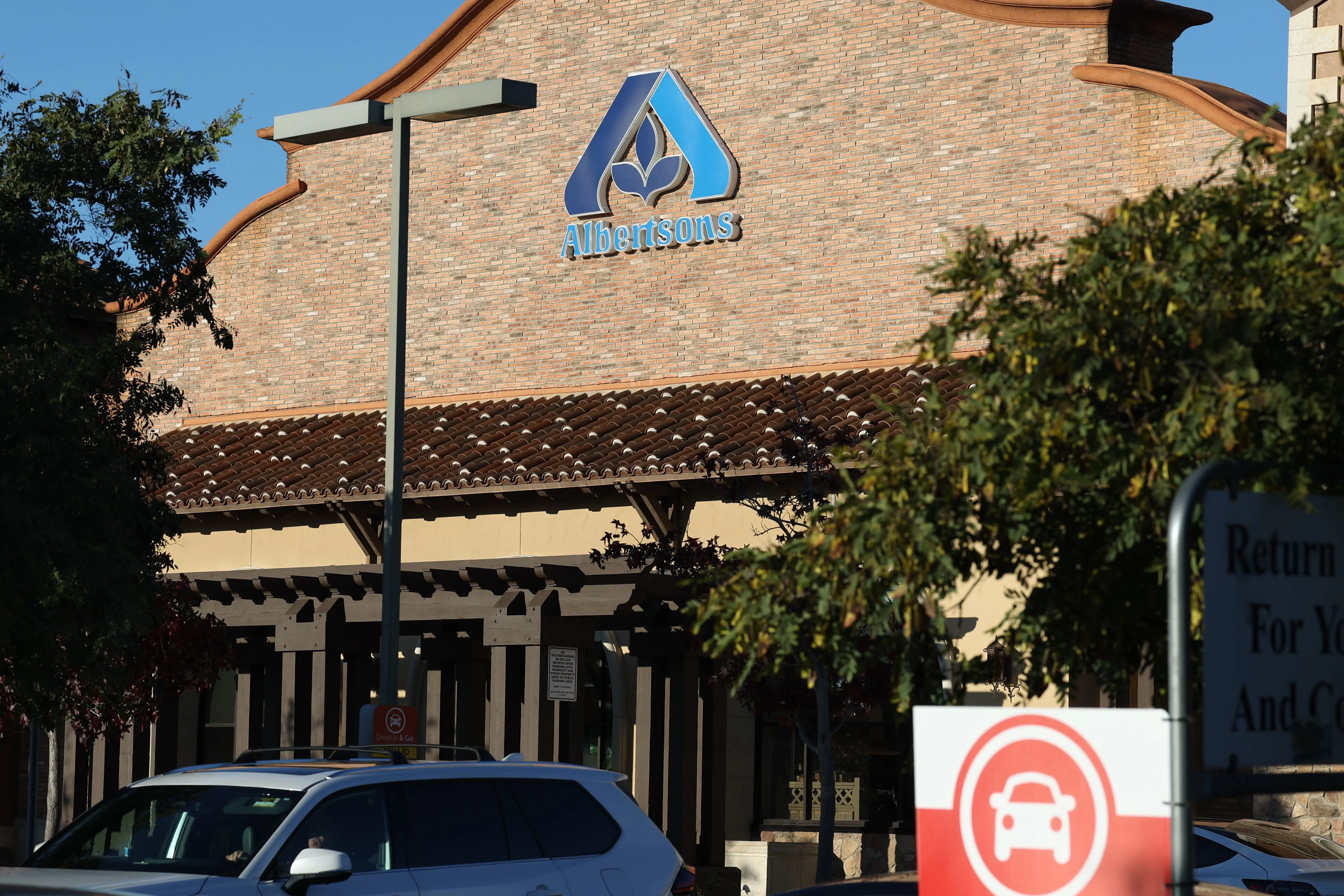 Albertsons storefront with logo above the entrance, parked cars, trees, and a Drive Up sign in front.