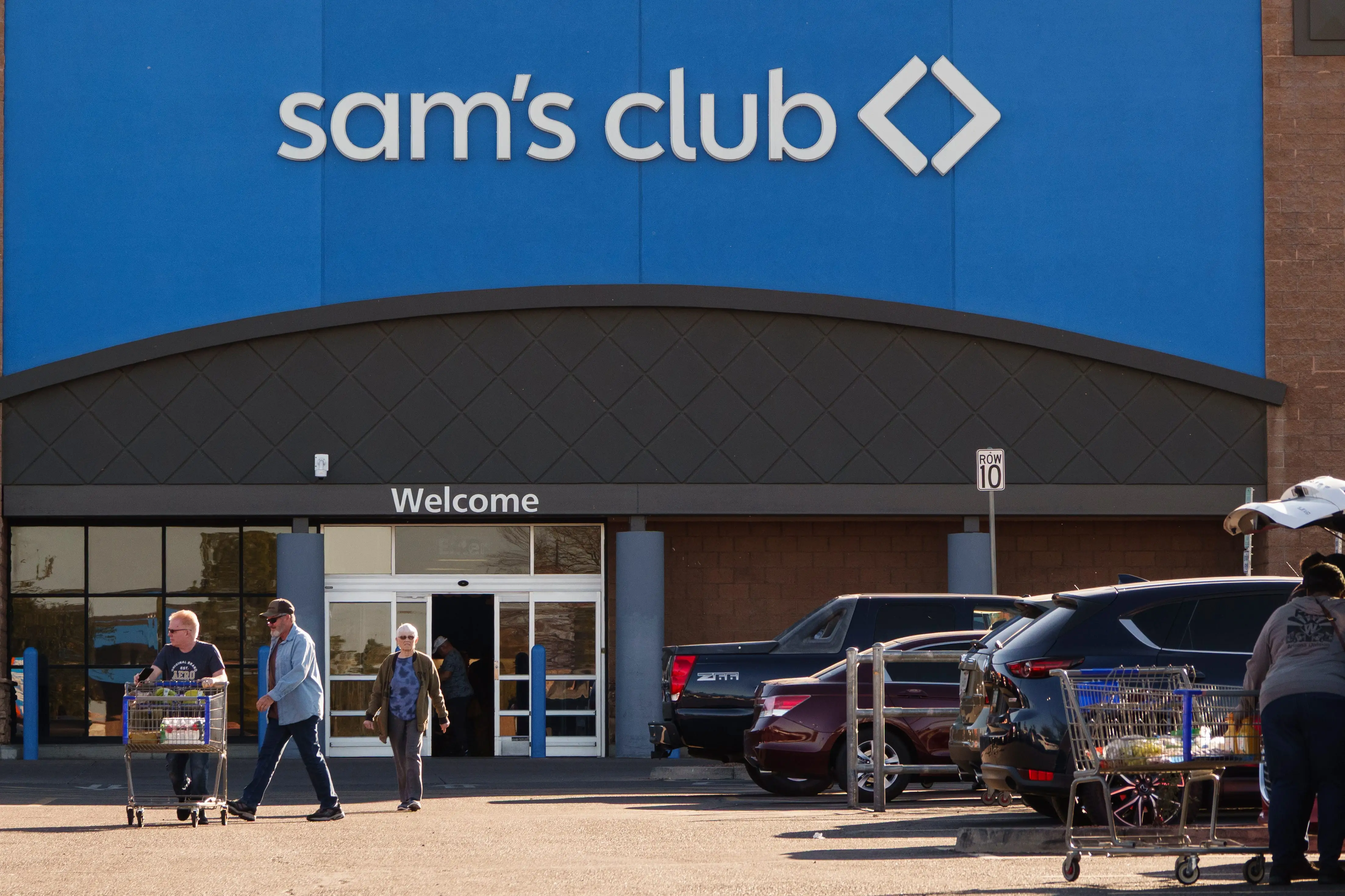 Sam's Club storefront with shoppers, carts, and parked vehicles outside the entrance.