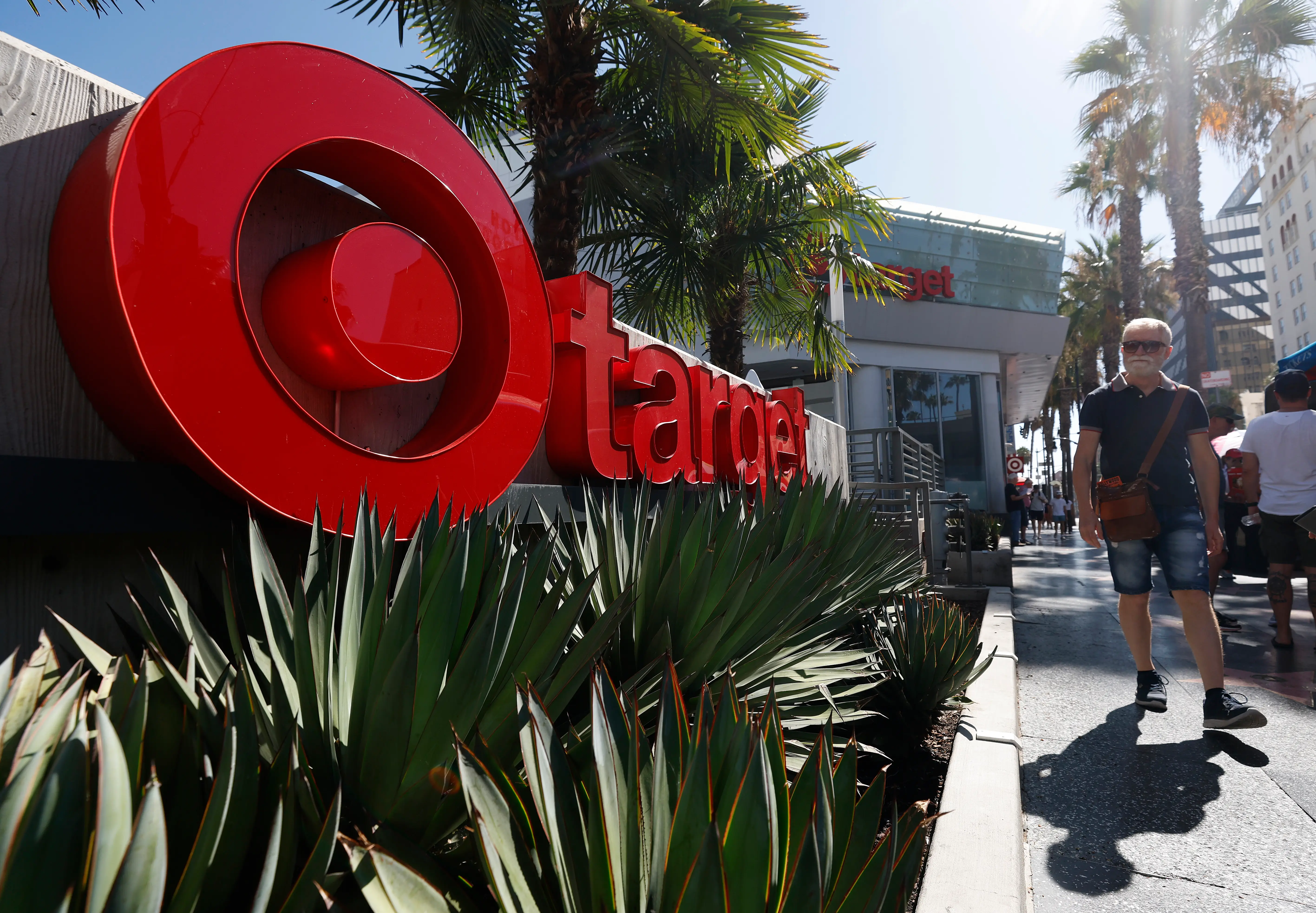 Large red Target logo and storefront sign stand beside palm trees and pedestrians on a sunny sidewalk.