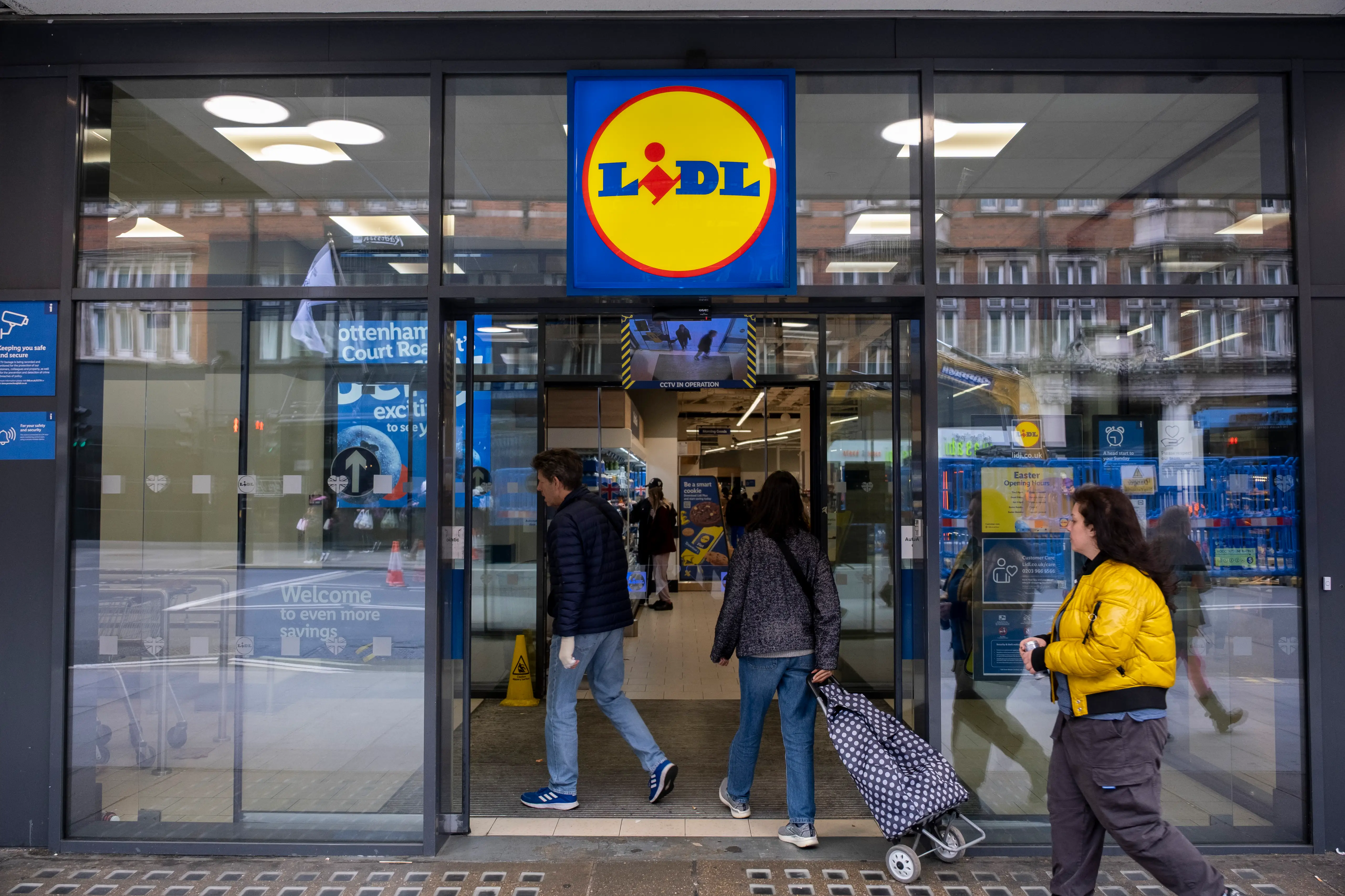 Lidl storefront with glass entrance doors, shoppers entering, and the yellow Lidl logo sign above the doorway.