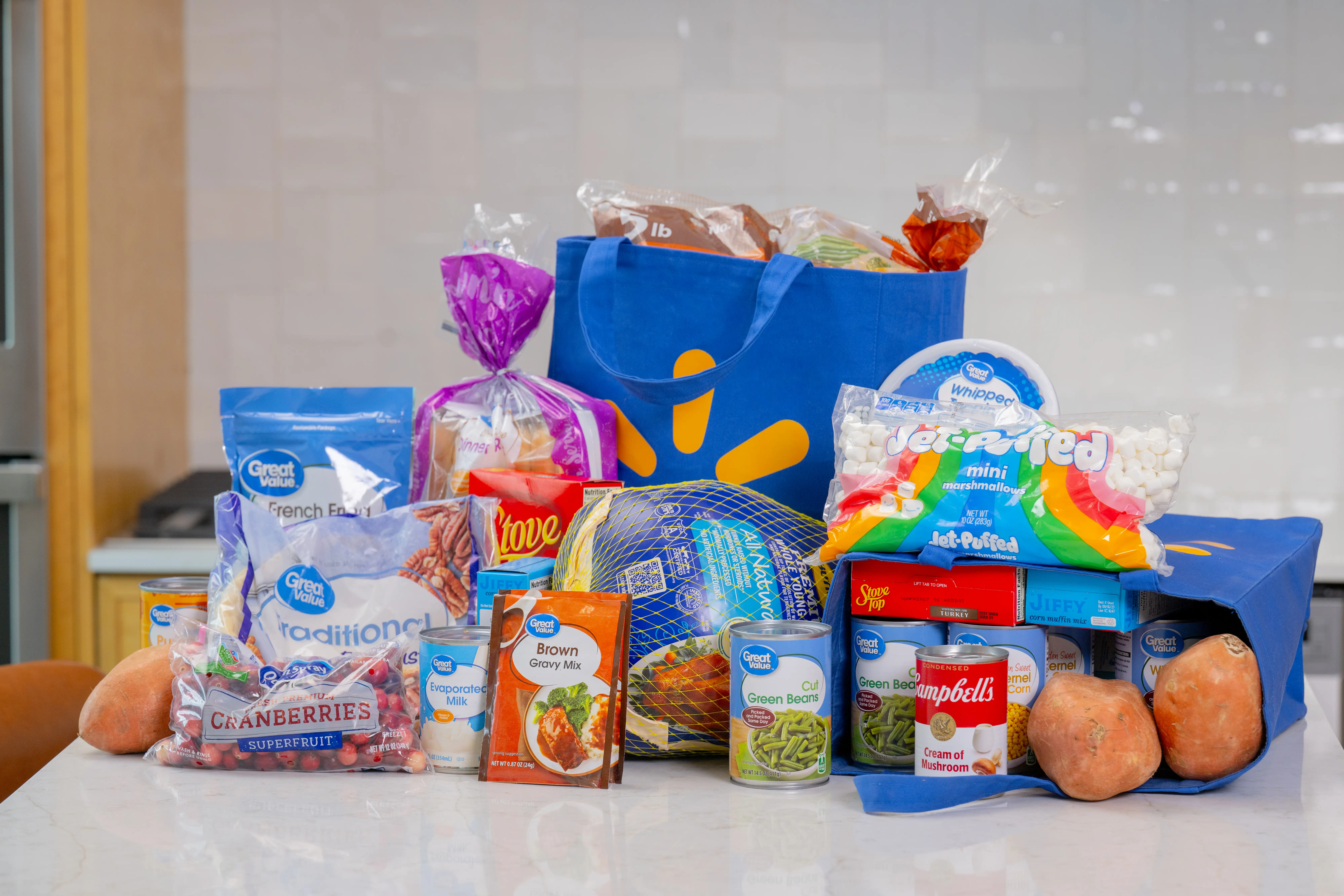 Assorted groceries including a turkey, canned goods, marshmallows, cranberries and sweet potatoes arranged beside blue reusable bags.