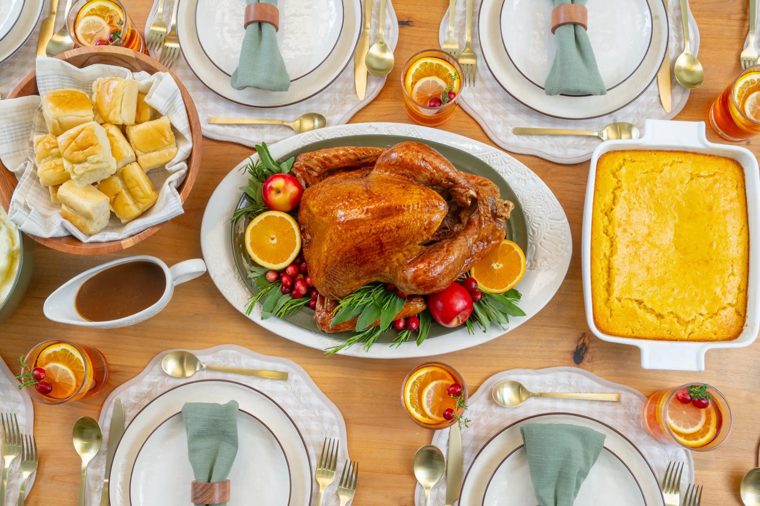 Overhead table setting with roasted turkey, rolls, gravy, cornbread, citrus drinks, and plated place settings.